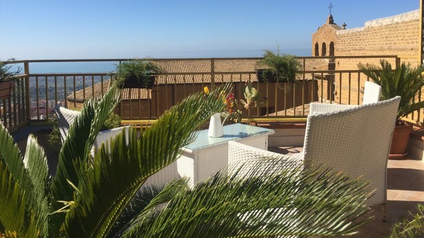 Une terrasse meublée de mobilier en osier, une table agrémentée d’un vase, et une vue sur un bâtiment doté d’un toit en tuiles et d’une tour.