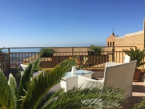 Une terrasse meublée de mobilier en osier, une table agrémentée d’un vase, et une vue sur un bâtiment doté d’un toit en tuiles et d’une tour.