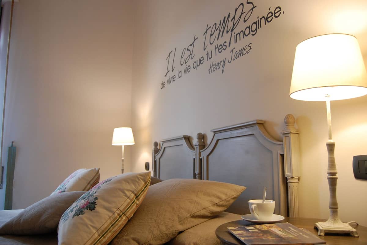A neatly made bed with floral pillows, a bedside table with a cup and saucer, and a wall with a quote.