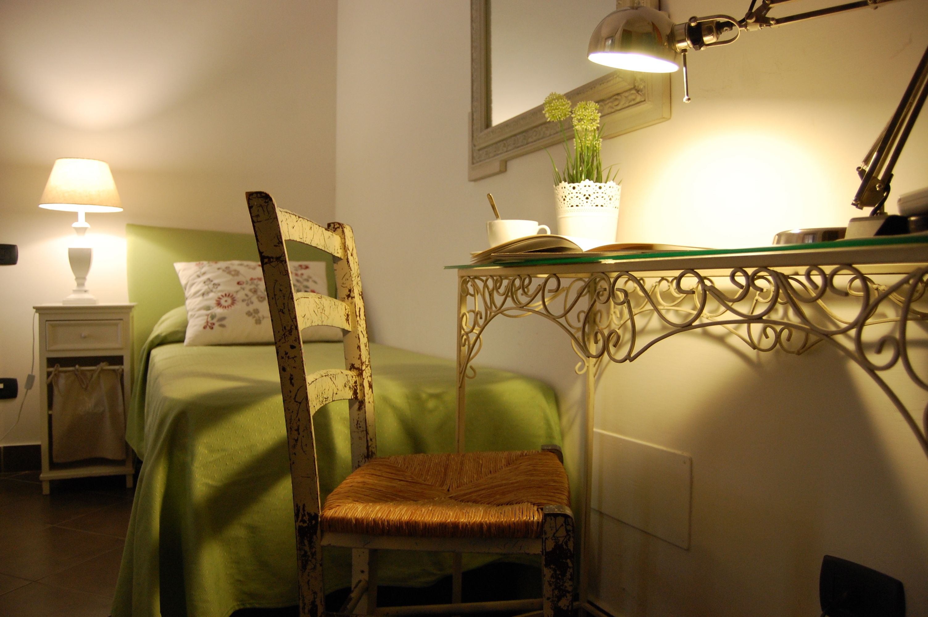 A bedroom with a green bed, a chair, a bedside table with a lamp, and a desk with a lamp and a plant.