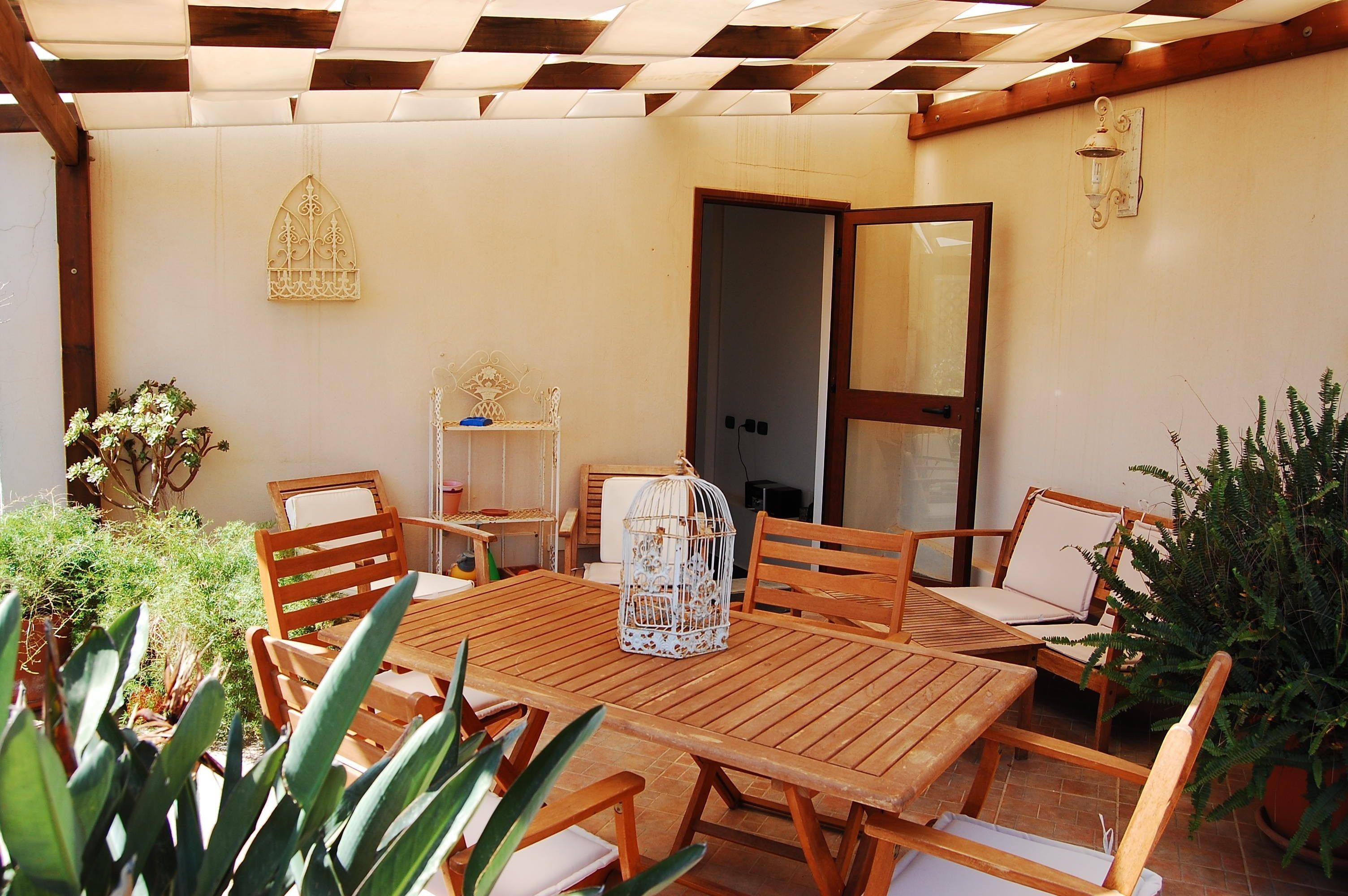 A wooden table and chairs set under a pergola, with a decorative birdcage on the table.