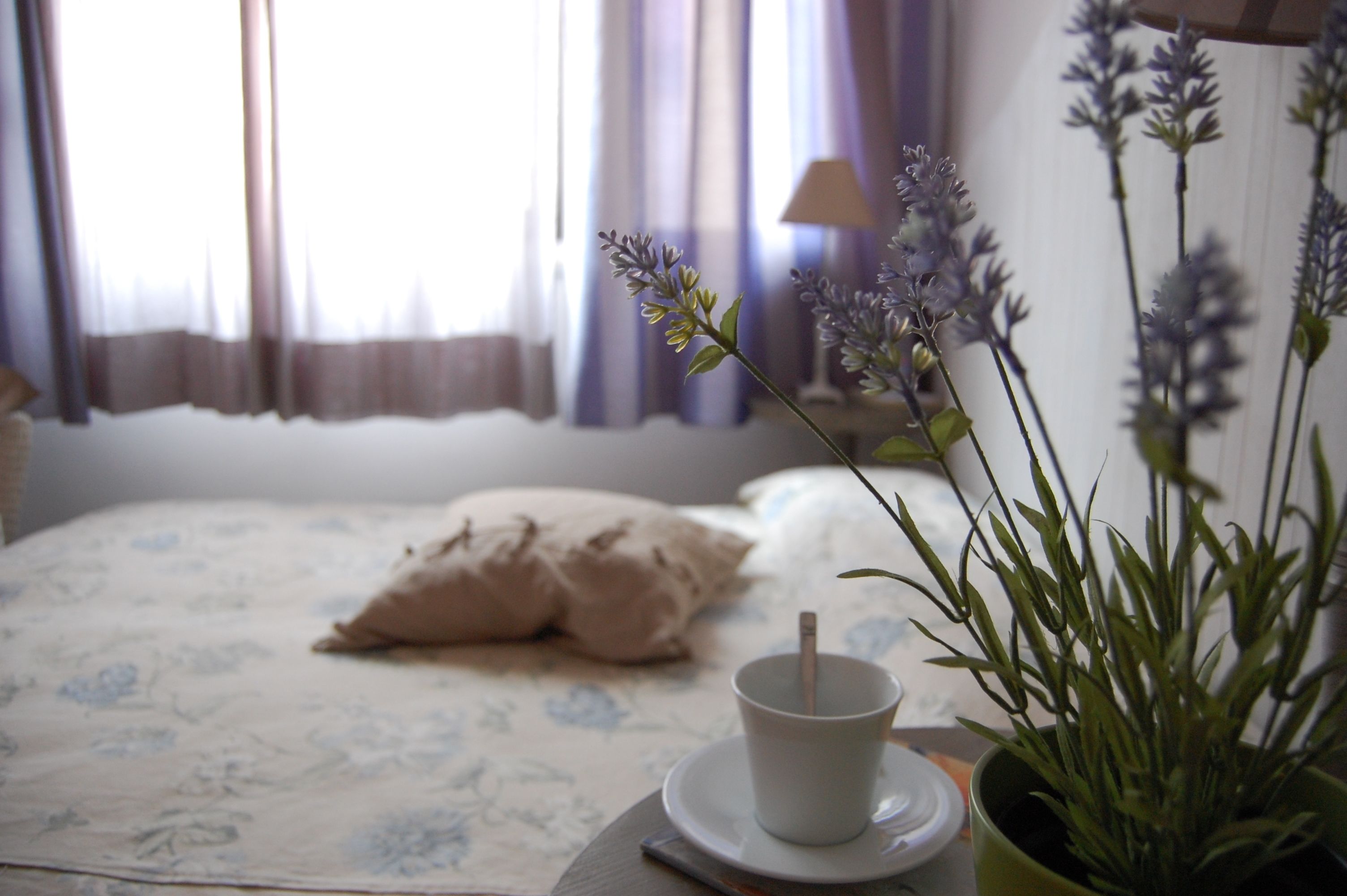 A neatly made bed with a floral-patterned bedspread, a white pillow, and a cup of tea on a bedside table.