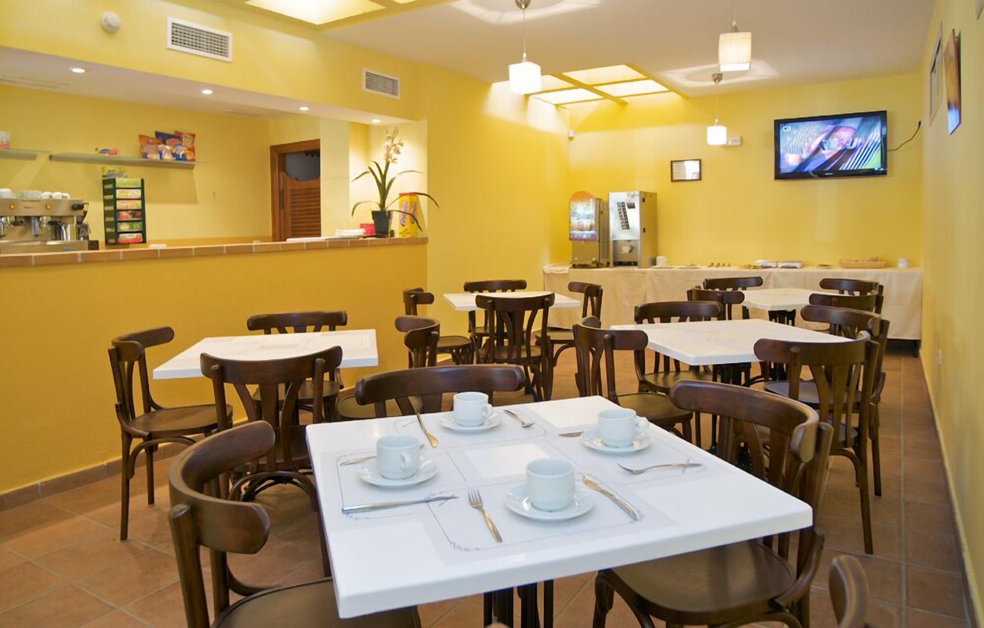 Un comedor con mesas dispuestas para cuatro, sillas, una barra con cafetera y un televisor en la pared.