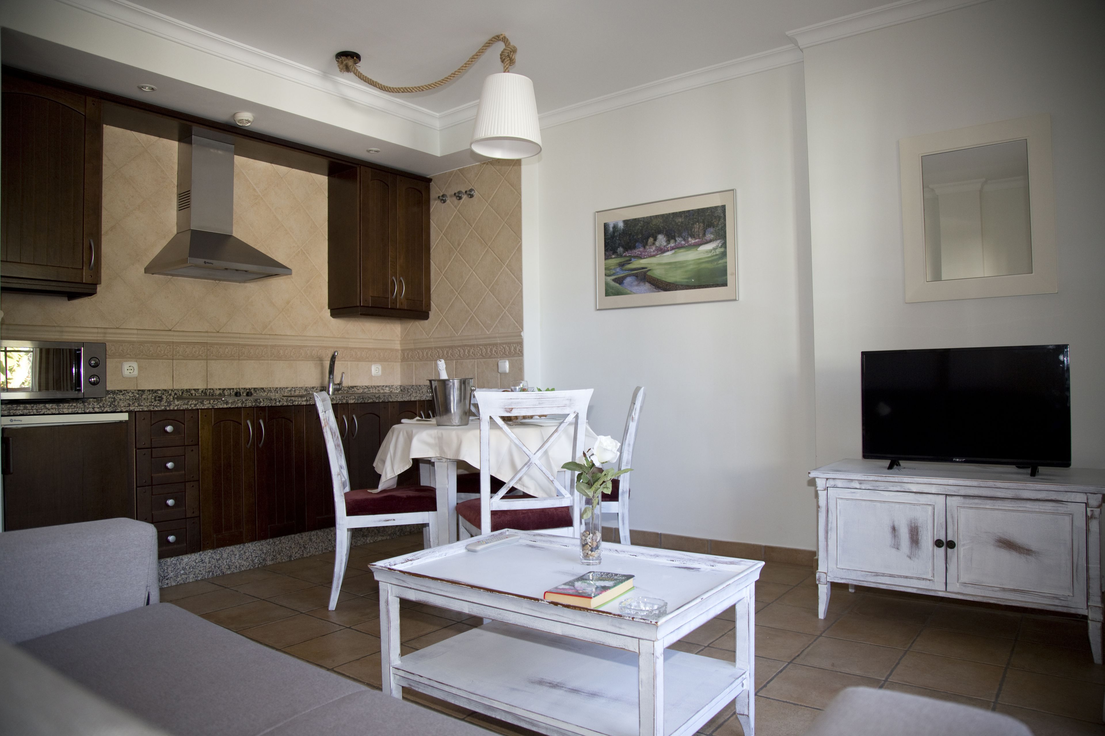 Una sala de estar con una mesa de centro blanca, un televisor sobre un mueble blanco, una mesa de comedor con sillas y una cocina con armarios de madera y una estufa.