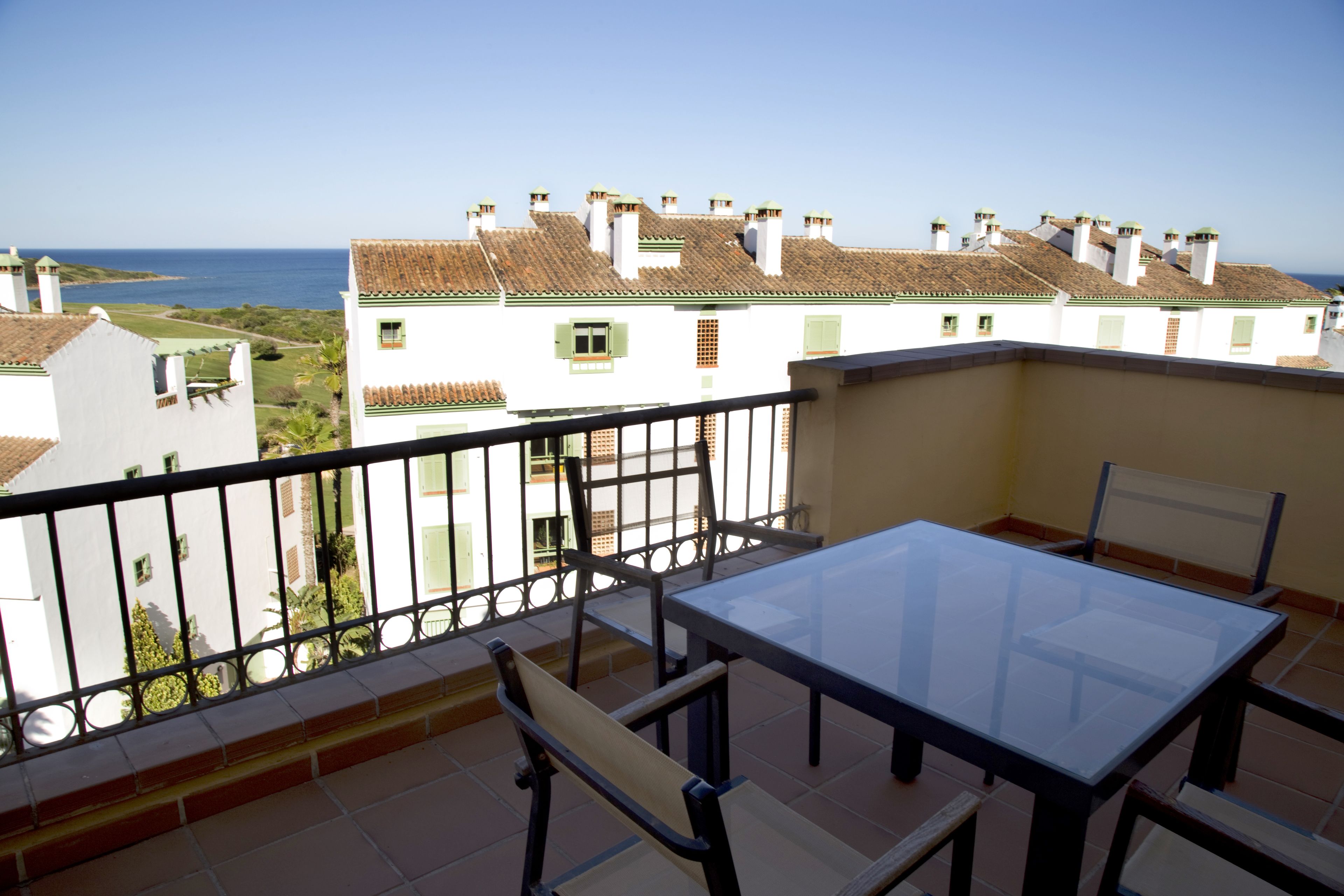 A balcony with a glass table and chairs, offering a view of the sea and neighboring buildings.