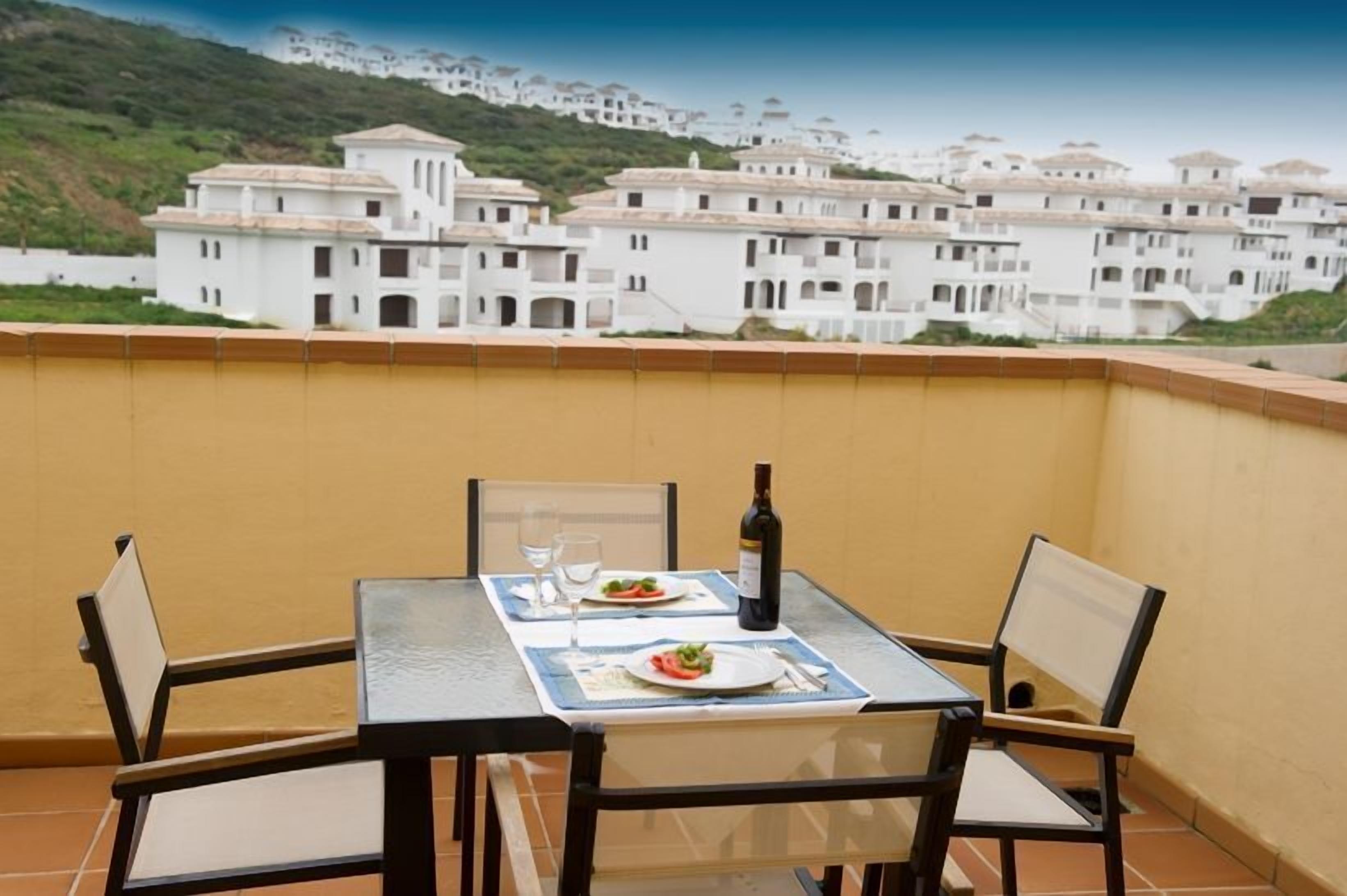 A balcony with a table set for two, a bottle of wine, and plates of food.