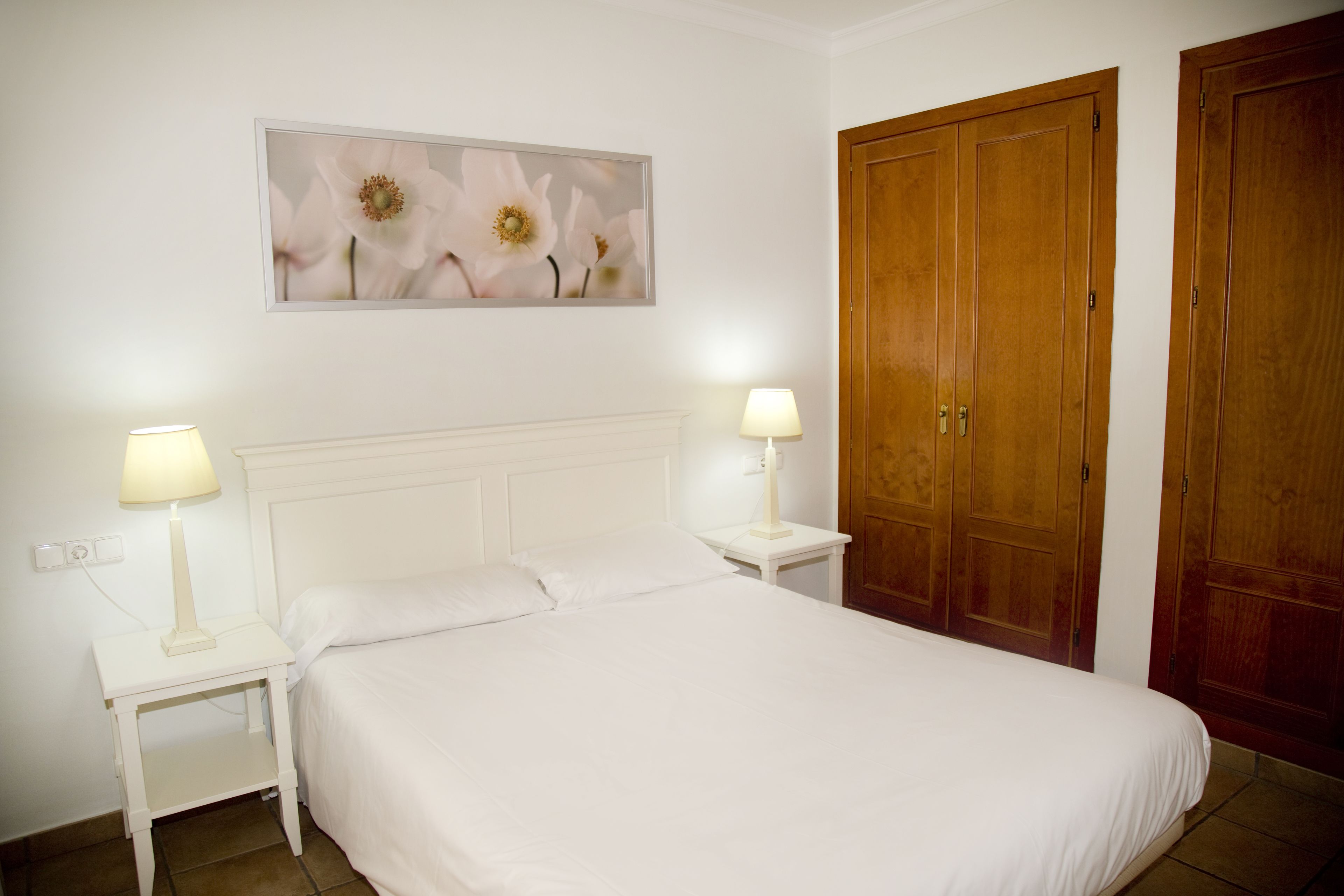 Un dormitorio con cama blanca, dos mesitas de noche con lámparas, un armario de madera y un cuadro floral en la pared.