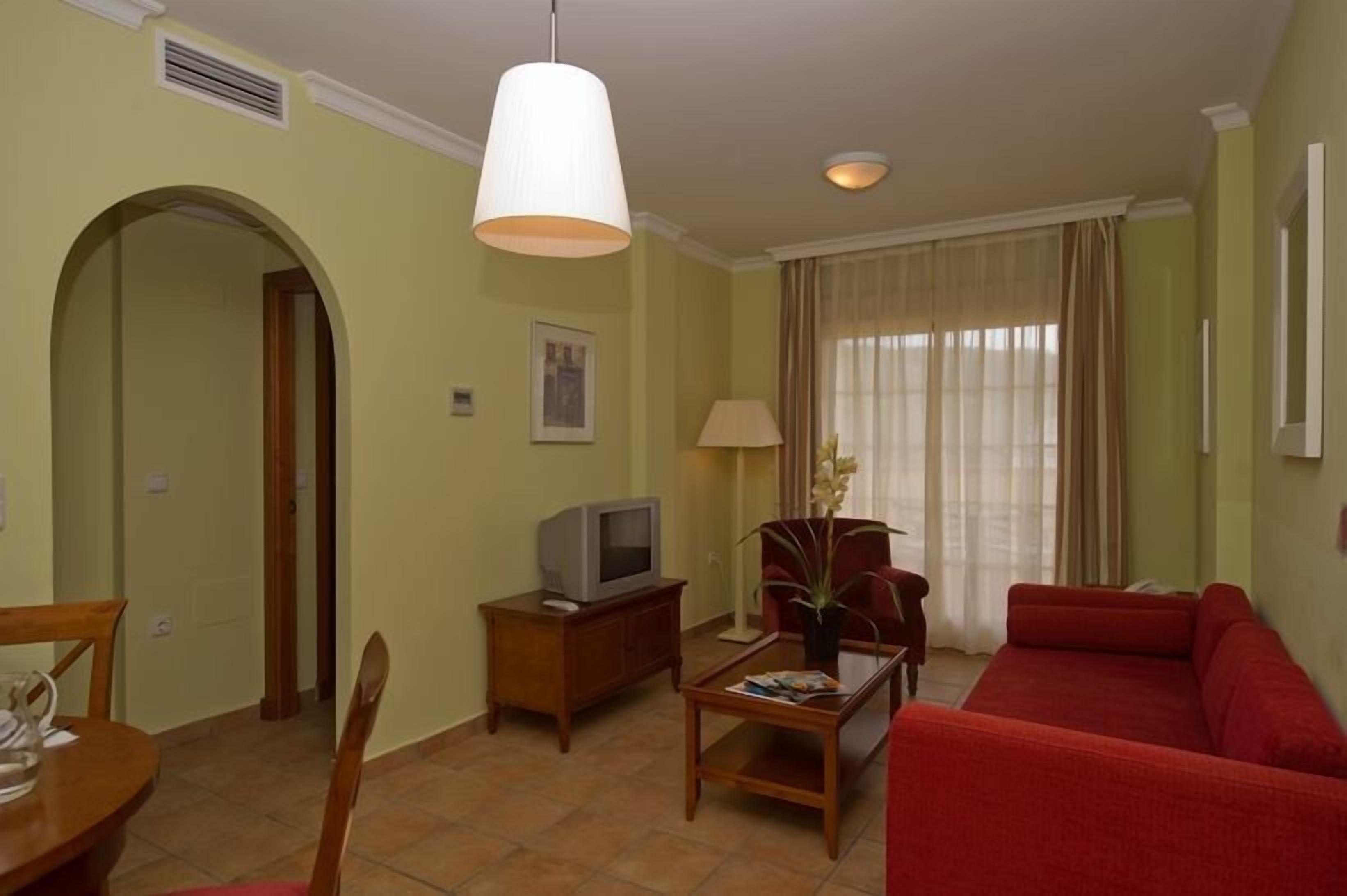 Una sala de estar con un sofá rojo, un televisor sobre un mueble de madera, una mesa con un florero y una lámpara.
