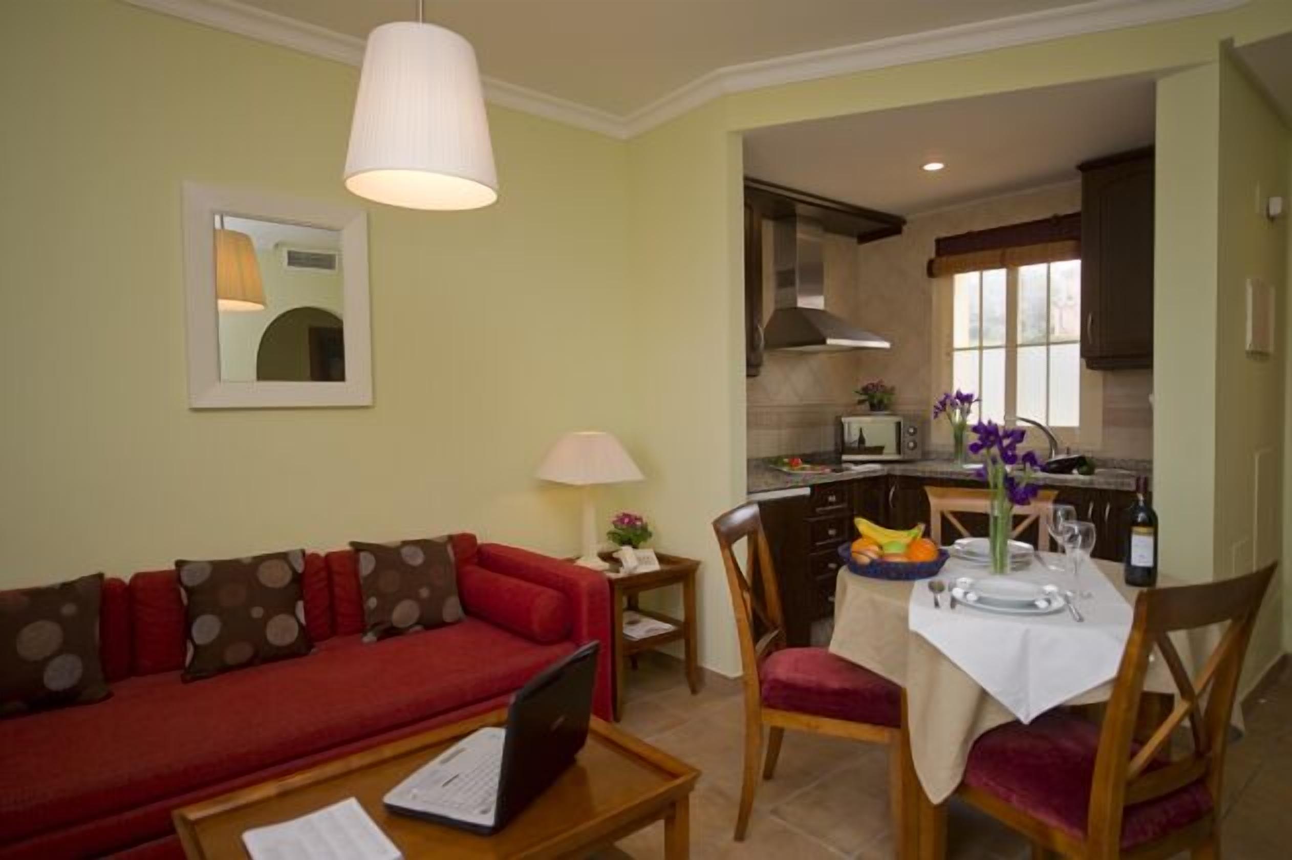 A compact living space with a red sofa, a dining table set for two, and a kitchenette in the background.
