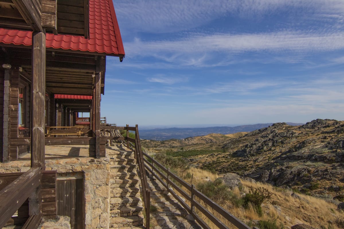 A wooden cabin with a red roof, stone steps, and a wooden railing overlooking a mountainous landscape.