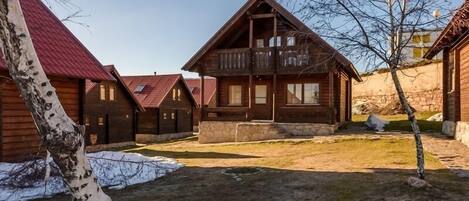 A wooden cabin with a balcony, surrounded by other cabins, a stone wall, and a birch tree.