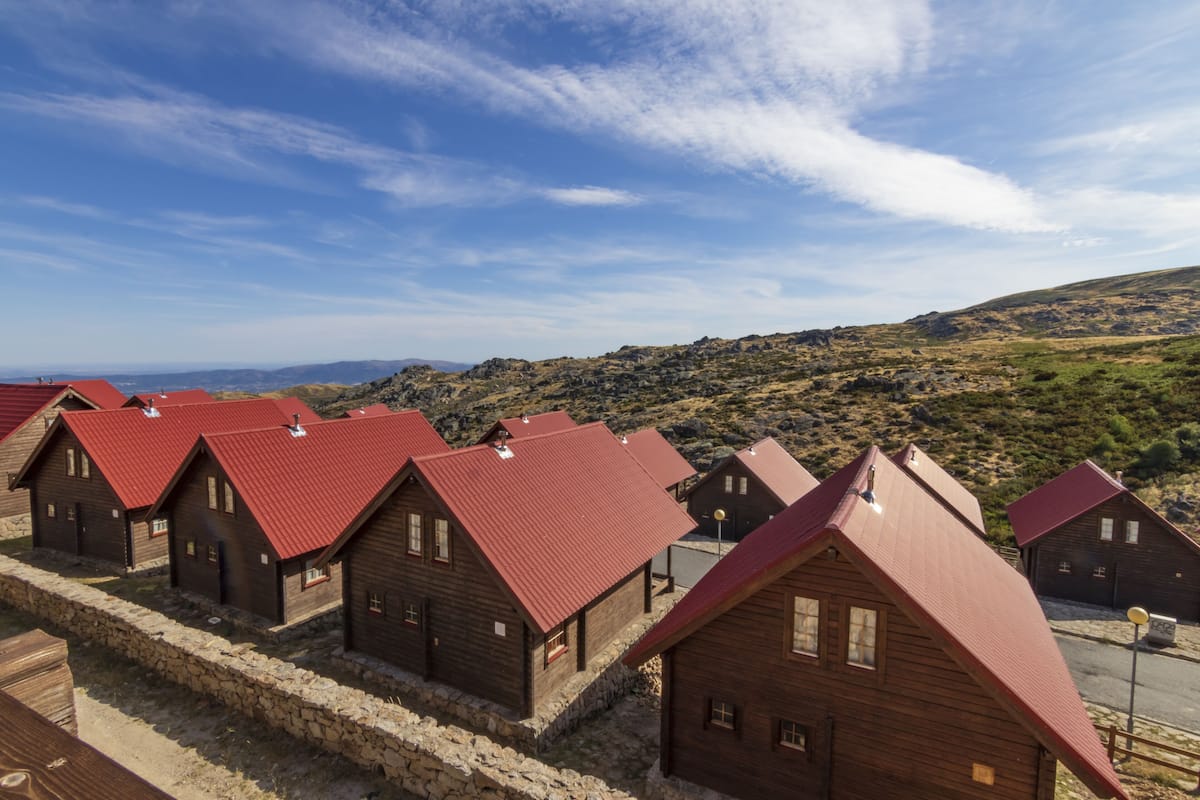 A row of wooden cabins with red roofs, arranged in a line along a stone wall, set against a hilly landscape under a clear sky.