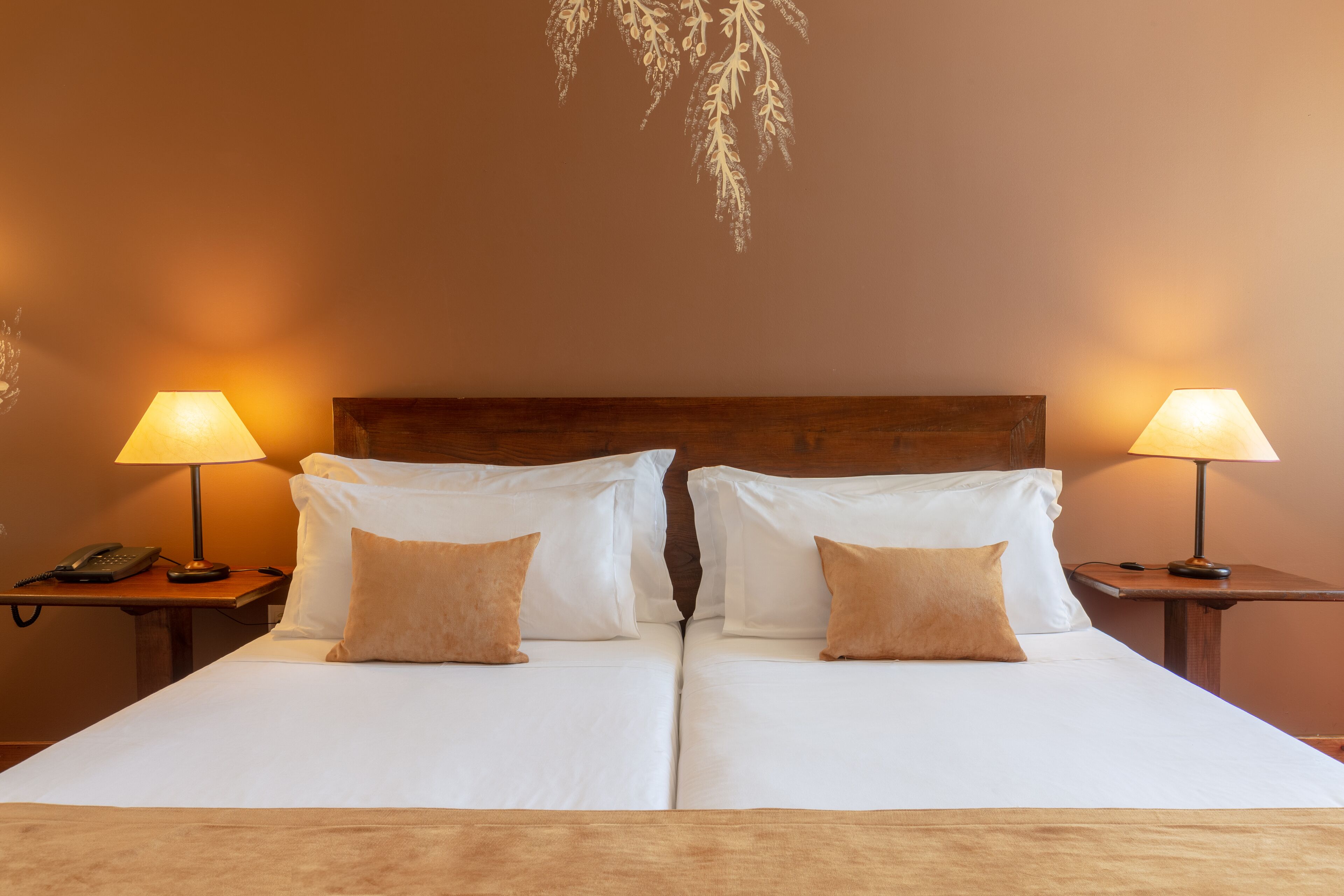 Una cama doble con sábanas blancas y dos almohadas marrones, con mesitas de noche a ambos lados, cada una con una lámpara y un teléfono.