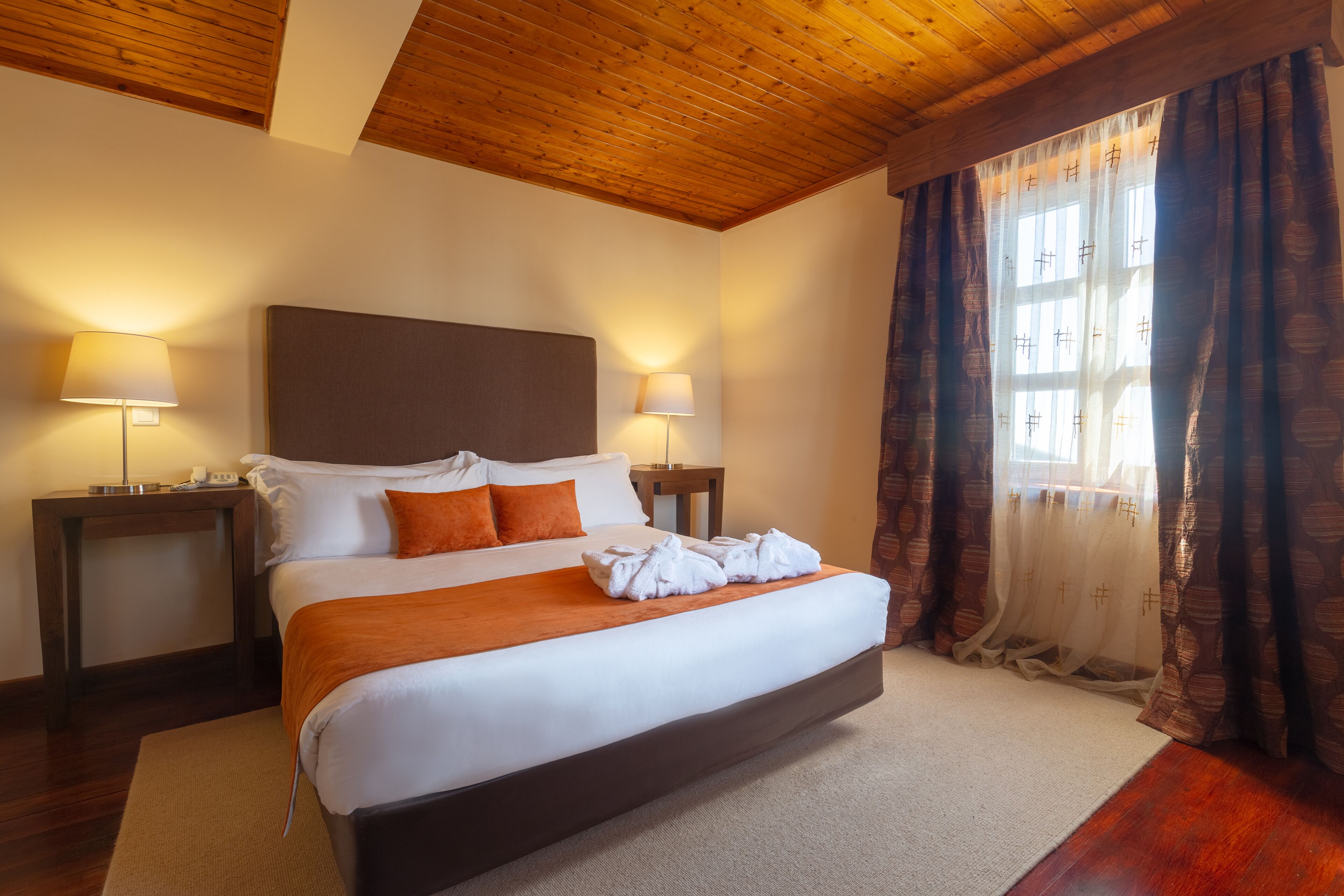 A large bed with a wooden headboard, two bedside tables with lamps, and a window with curtains.