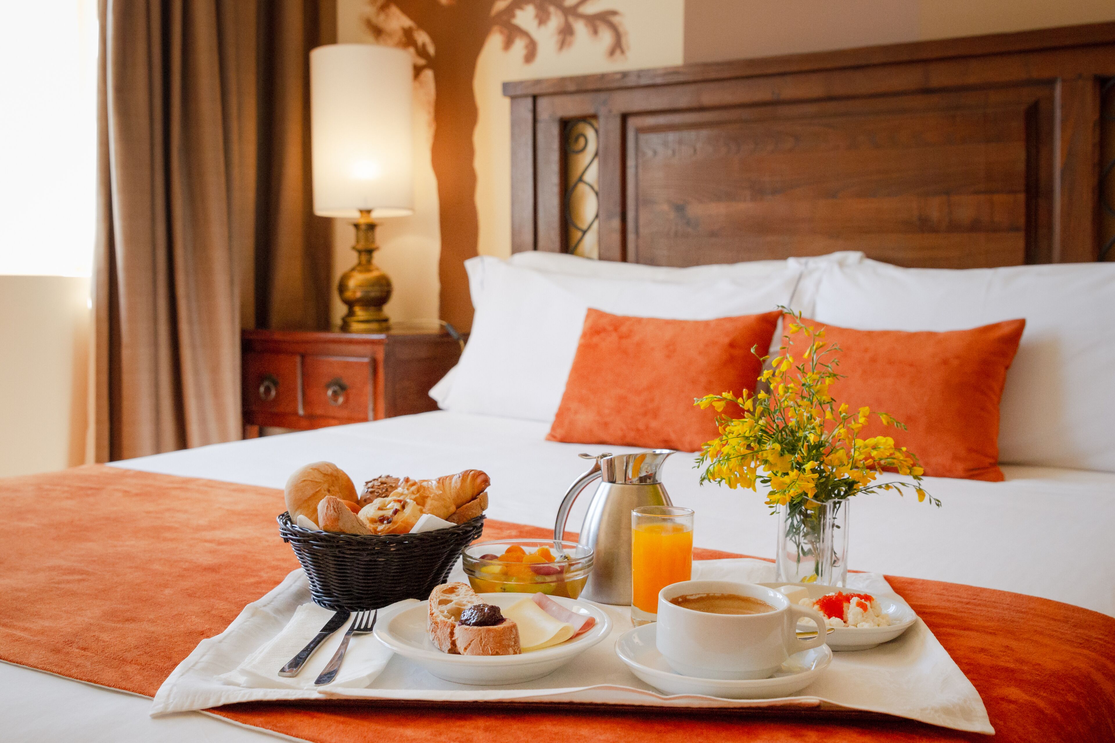A neatly made breakfast tray with croissants, fruits, and coffee on a bed with orange bedding and pillows.