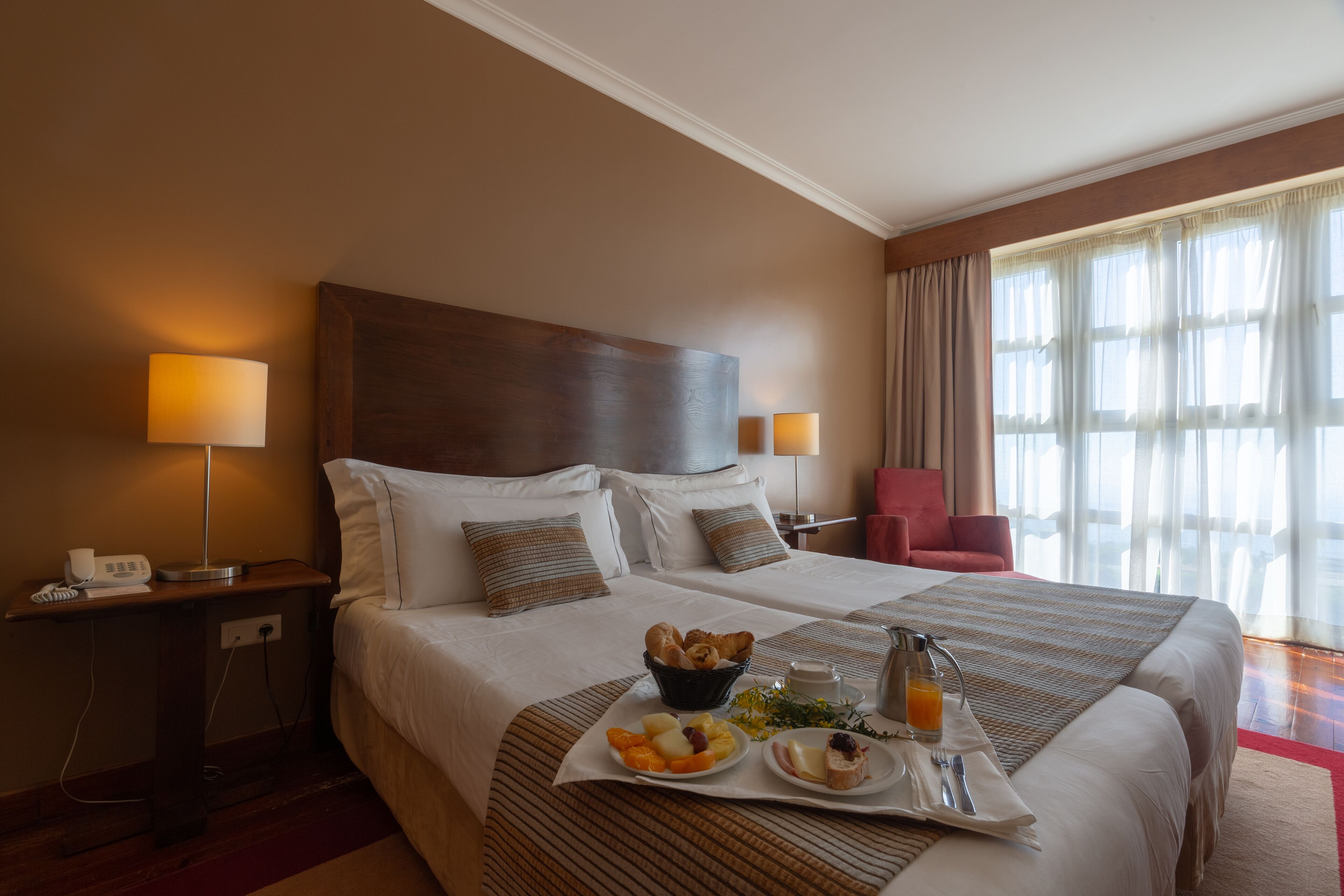 Ein Hotelzimmer mit einem großen Bett, Nachttischlampen, einem kleinen Schreibtisch und einem Frühstücksbrett mit Früchten und Gebäck.