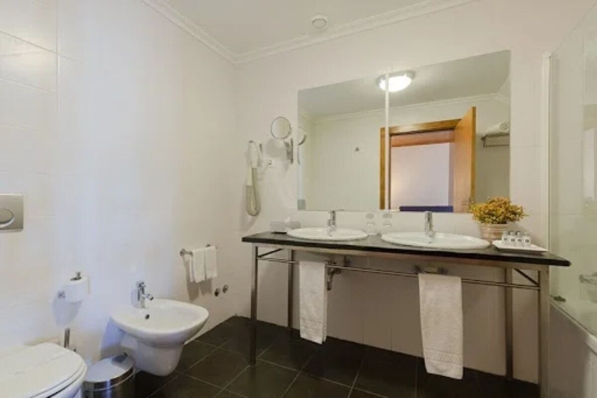 Een badkamer met een hygiëneschuiver, toilet en een dubbele wastafel met spiegel erboven.