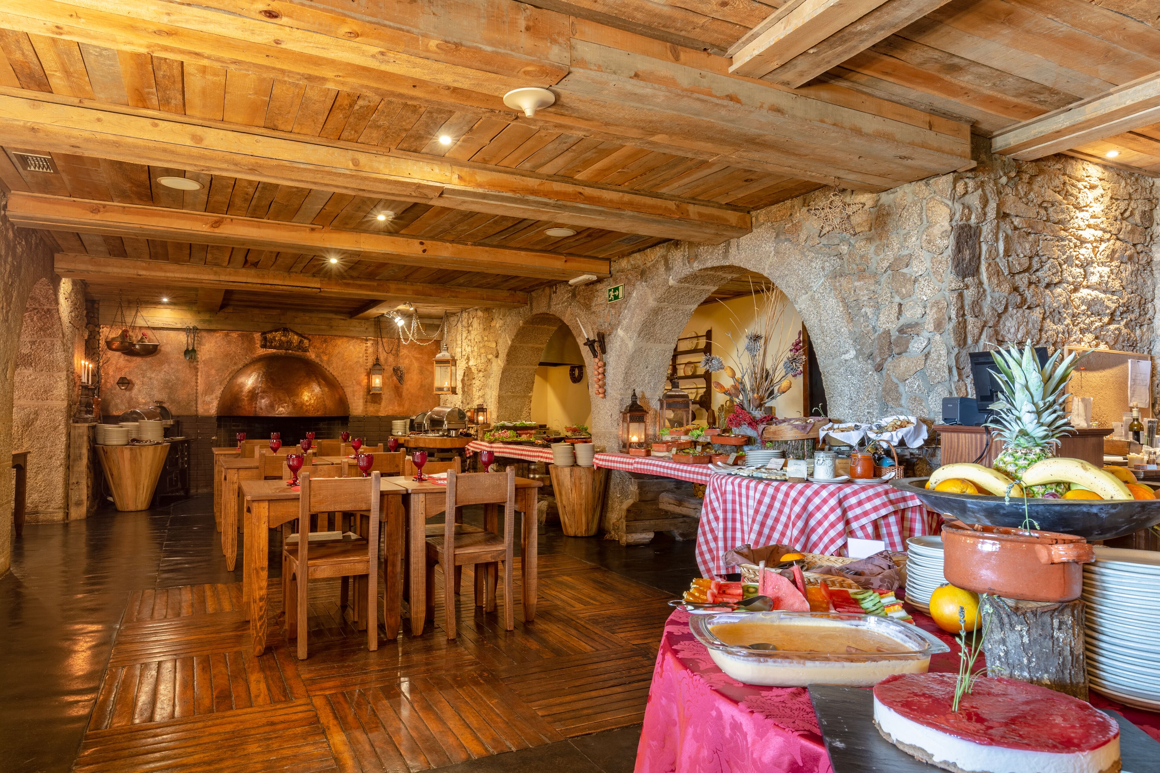A rustic dining area with wooden tables and chairs, a stone wall, and a variety of food and drink items on a table.