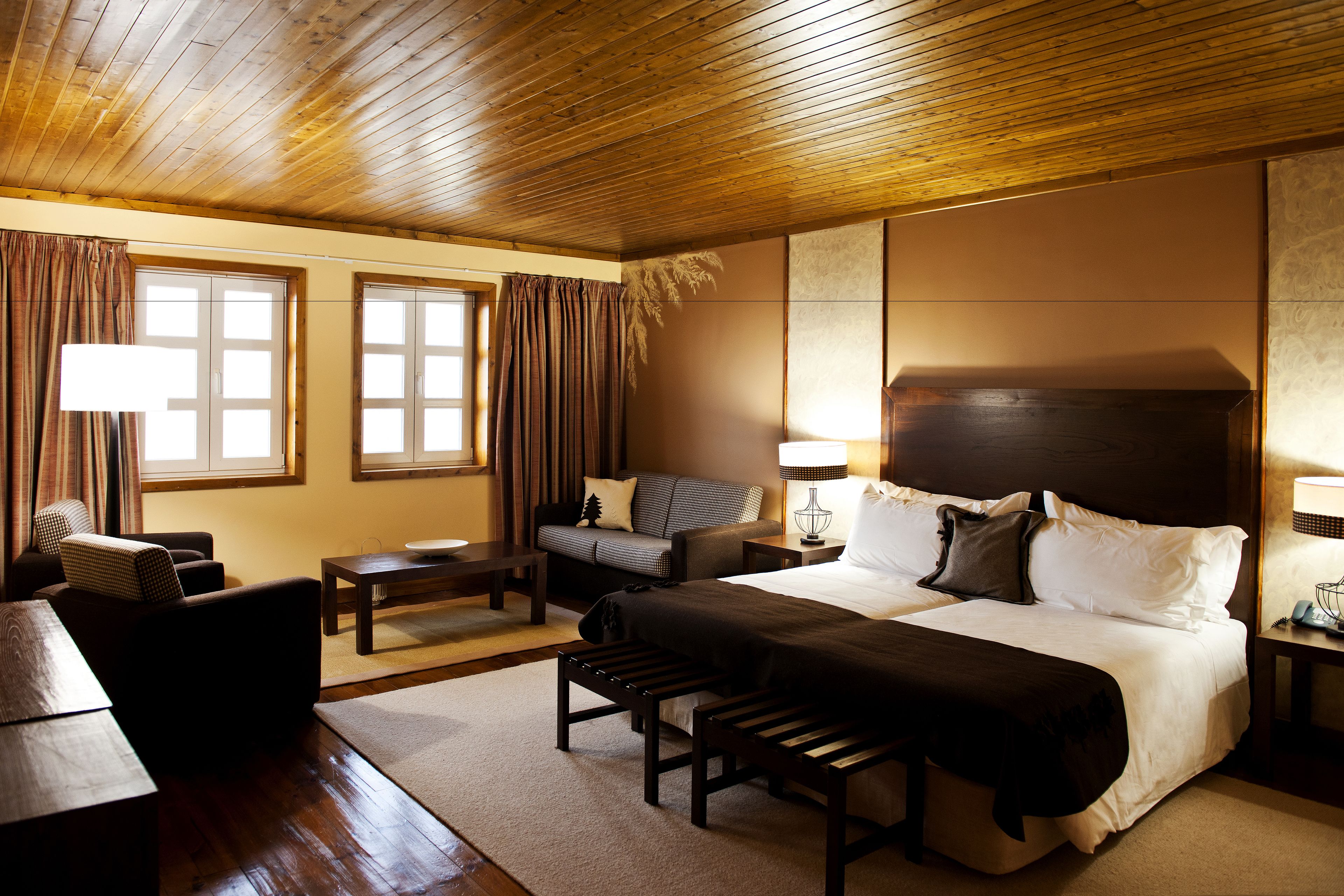 A hotel room with a wooden ceiling, a large bed with white linens, a sofa, a desk, and two lamps.