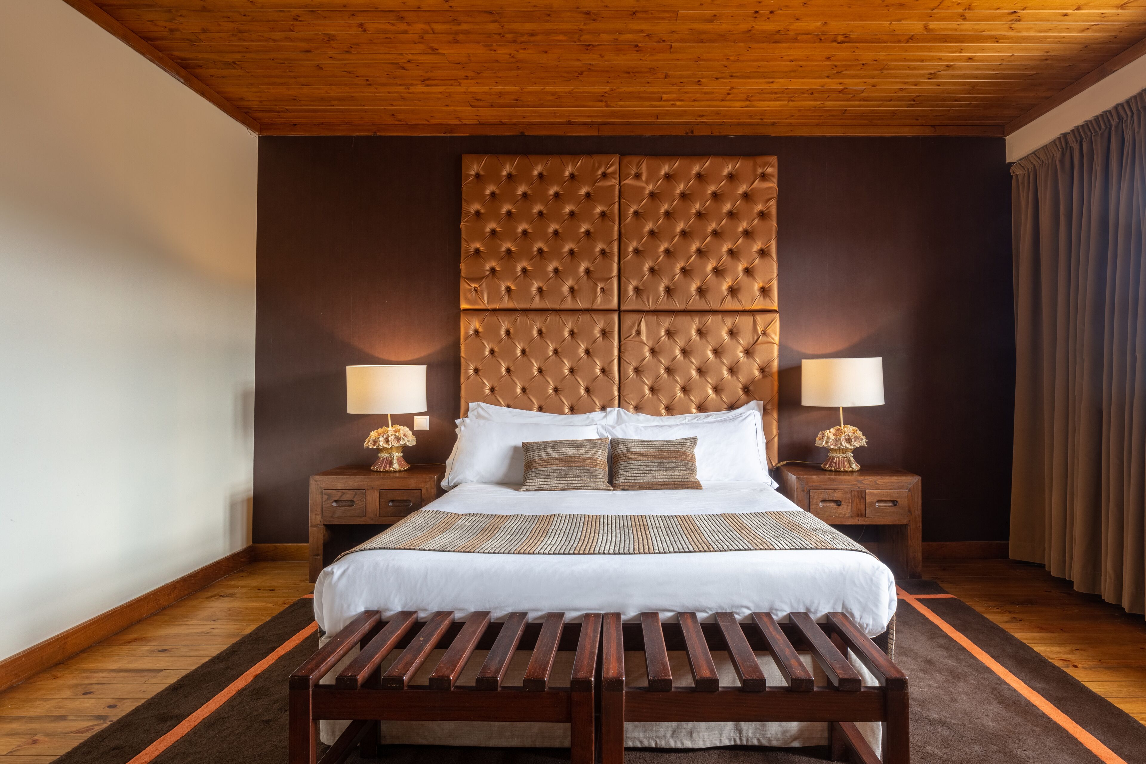 Un dormitorio ordenado con una cama grande, dos mesitas de noche con lámparas, un banco de madera y un cabecero capitonado.