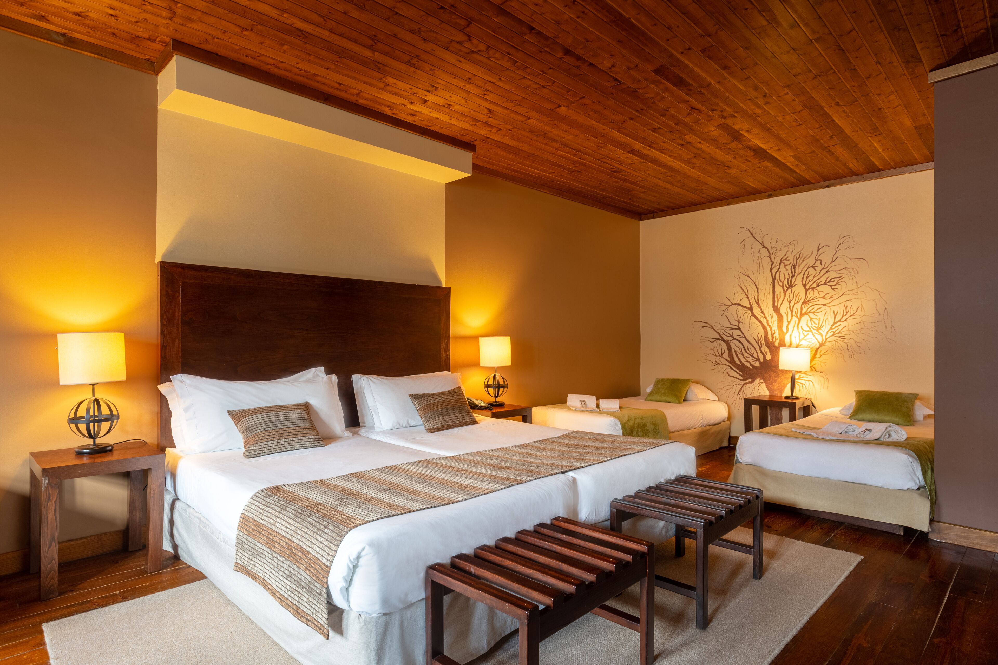 A hotel room with a large bed, two smaller beds, wooden headboard, bedside lamps, and a decorative tree wall art.