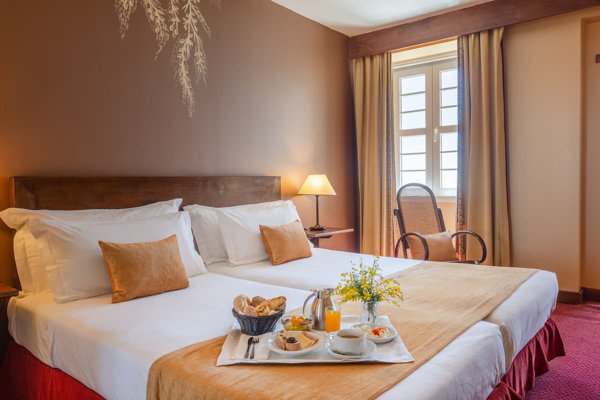 Een hotelkamer met een net opgemaakt bed, een ontbijttafel met gebak, een koffiezetapparaat en een vaas met bloemen.