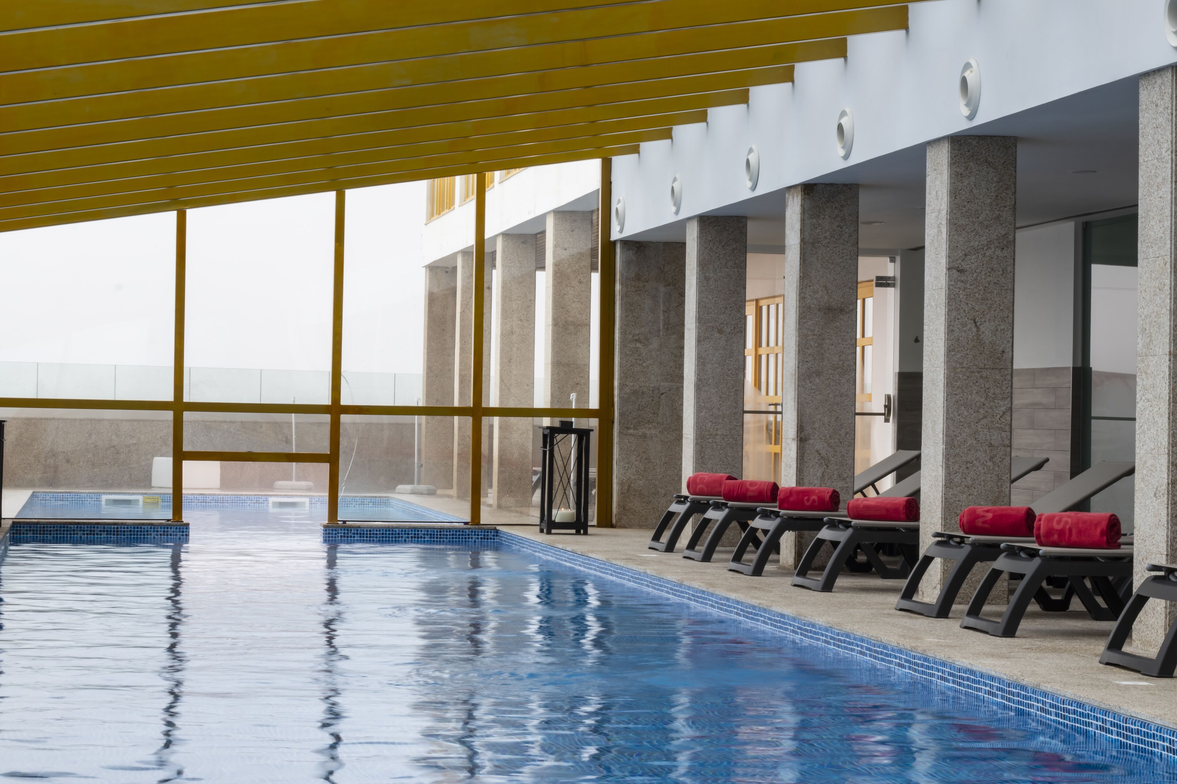 Una piscina con agua azul cristalina, rodeada de columnas y toallas rojas en sillas de descanso.