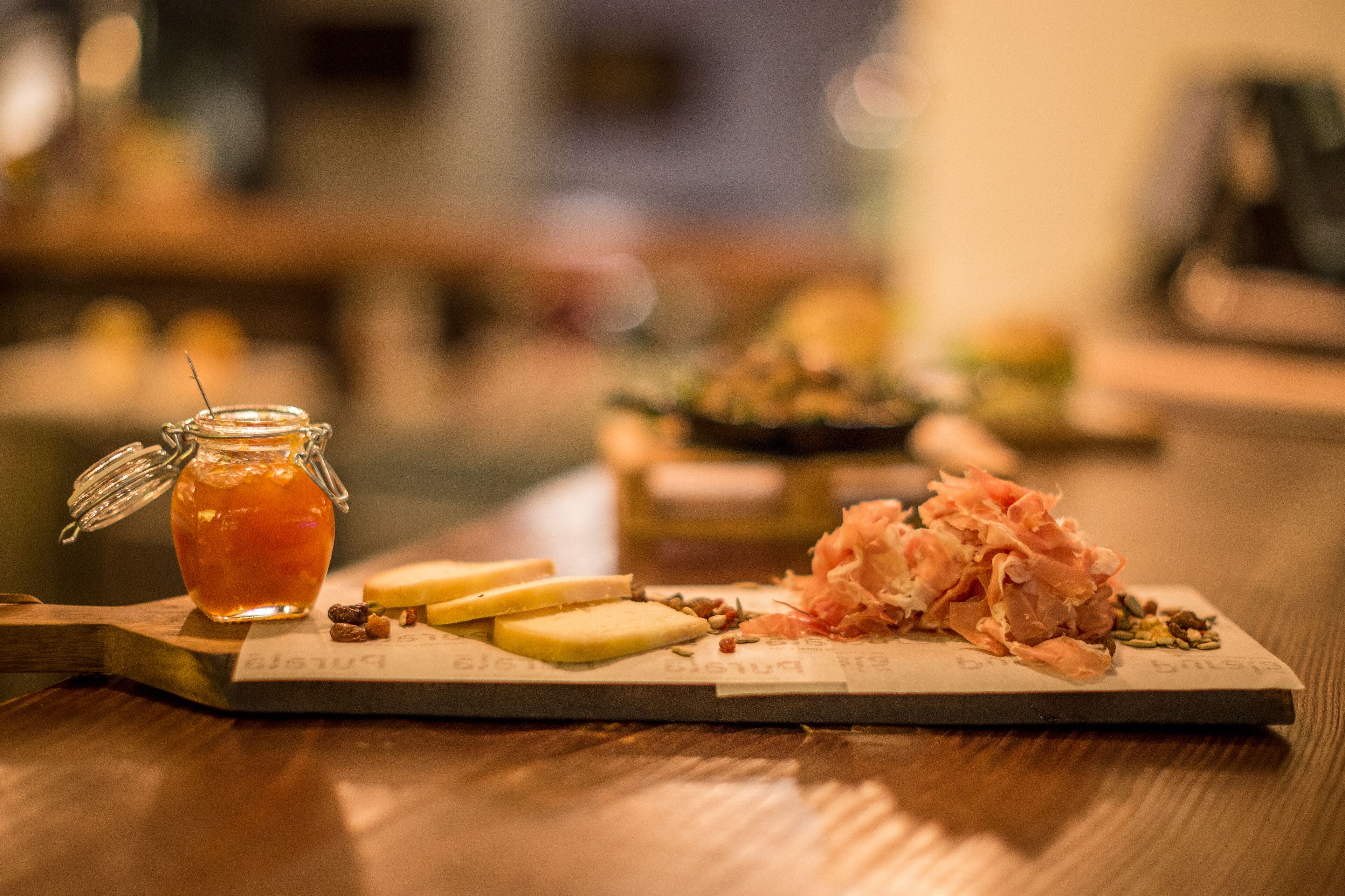 A wooden table with a glass jar of honey, cheese slices, and a plate of cured meats.