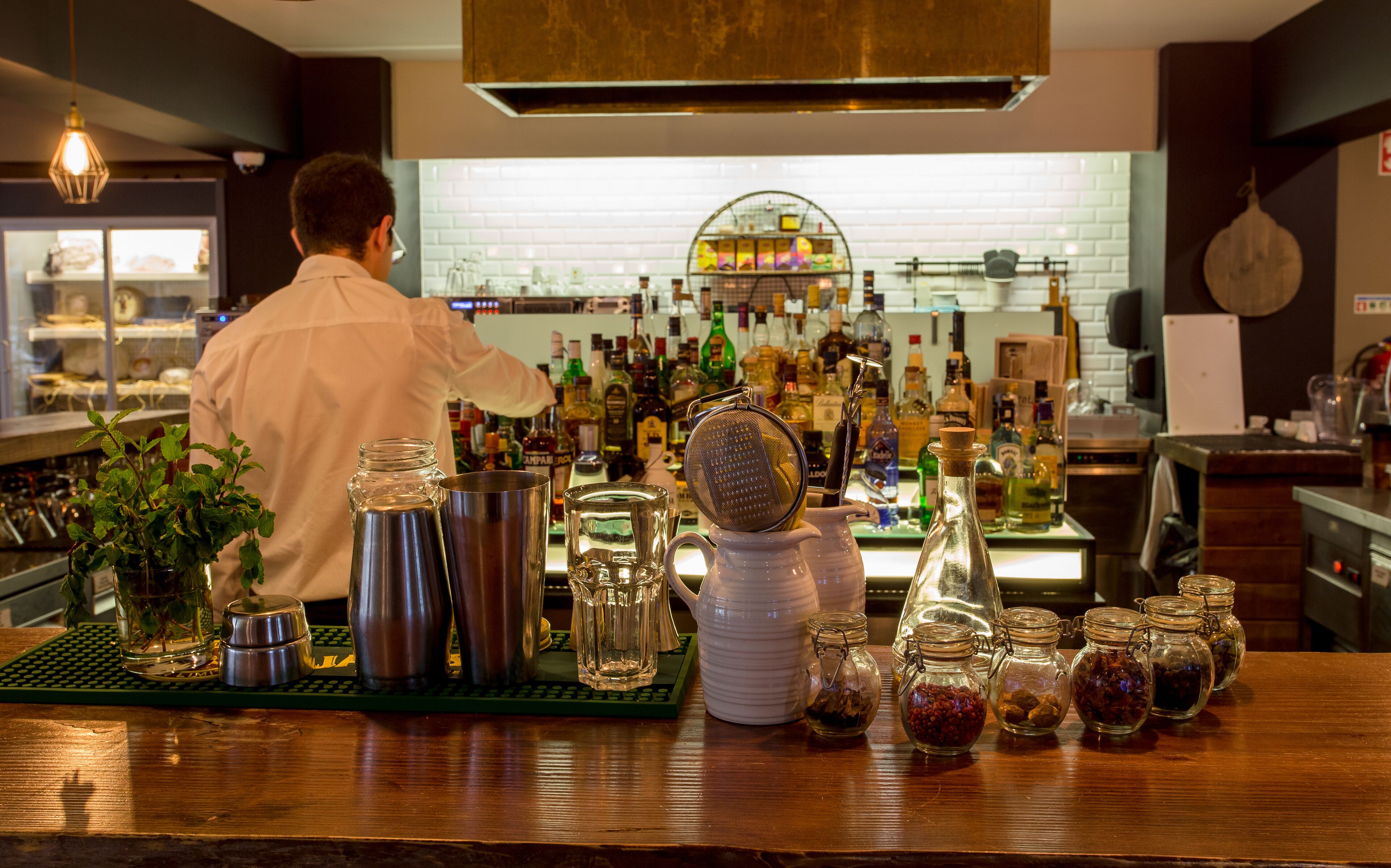 A bar area with a bartender preparing drinks, various bottles, and jars of spices on the counter.
