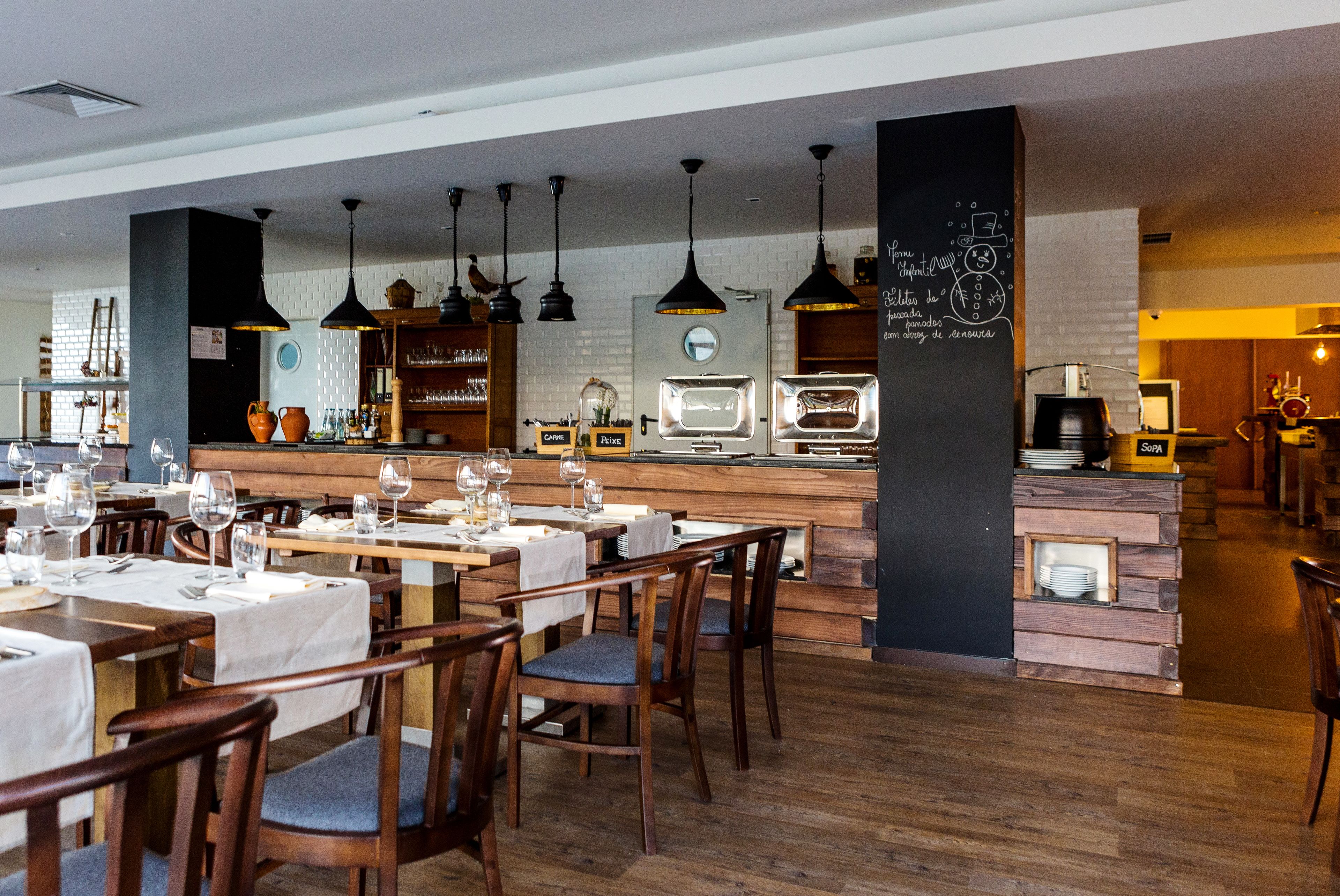 Un restaurante con mesas y sillas de madera, iluminación colgante y un menú en una pizarra.