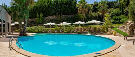 A swimming pool with lounge chairs and umbrellas, surrounded by greenery and a stone wall.
