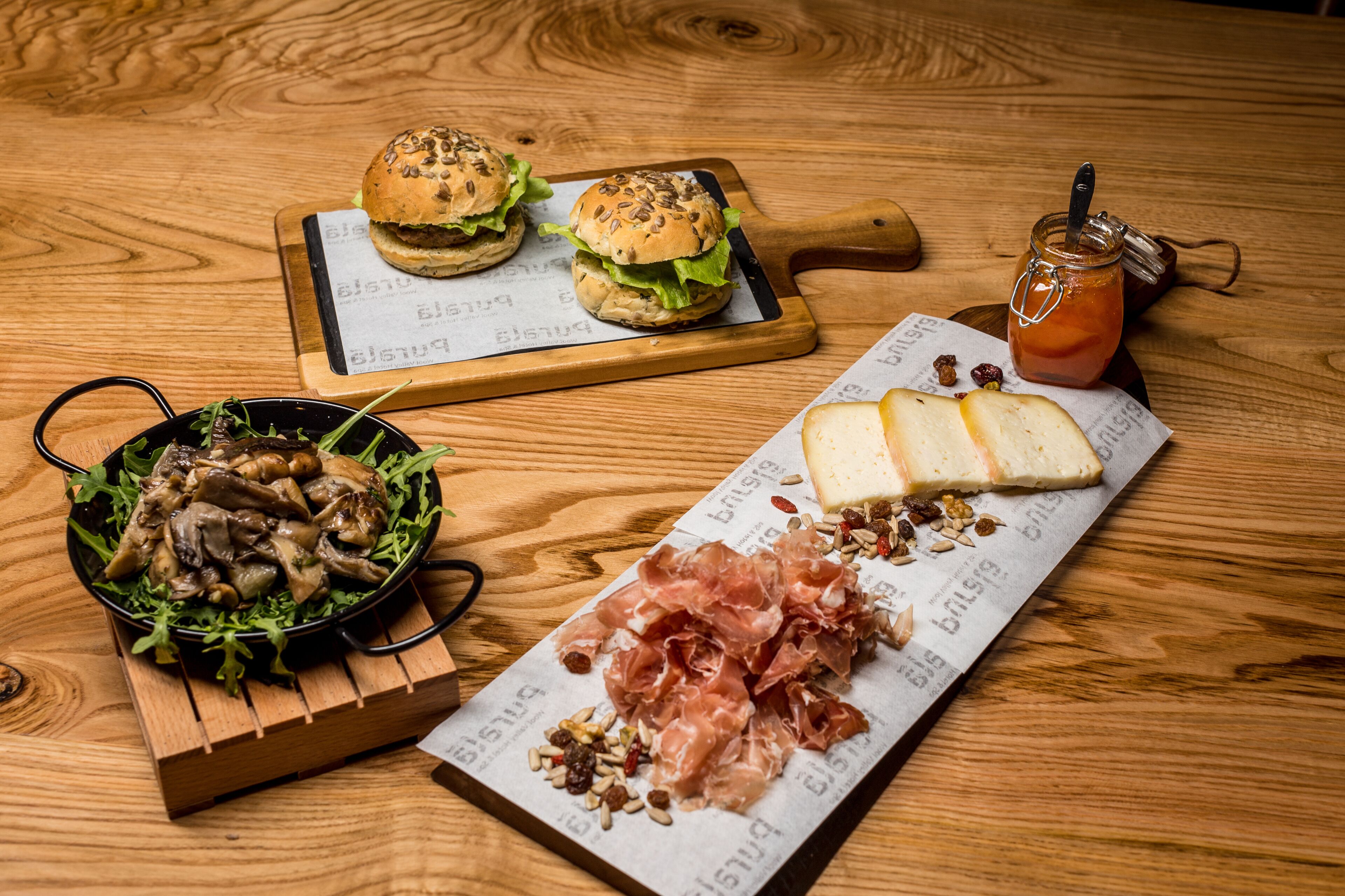 A wooden table with a burger, a bowl of mushrooms, a plate of cheese and prosciutto, and a jar of jam.