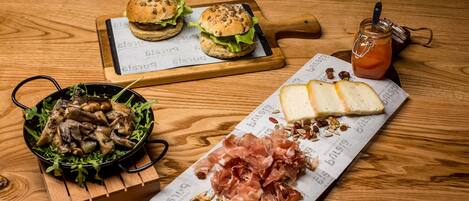 A wooden table with a burger, a bowl of mushrooms, a plate of cheese and prosciutto, and a jar of jam.