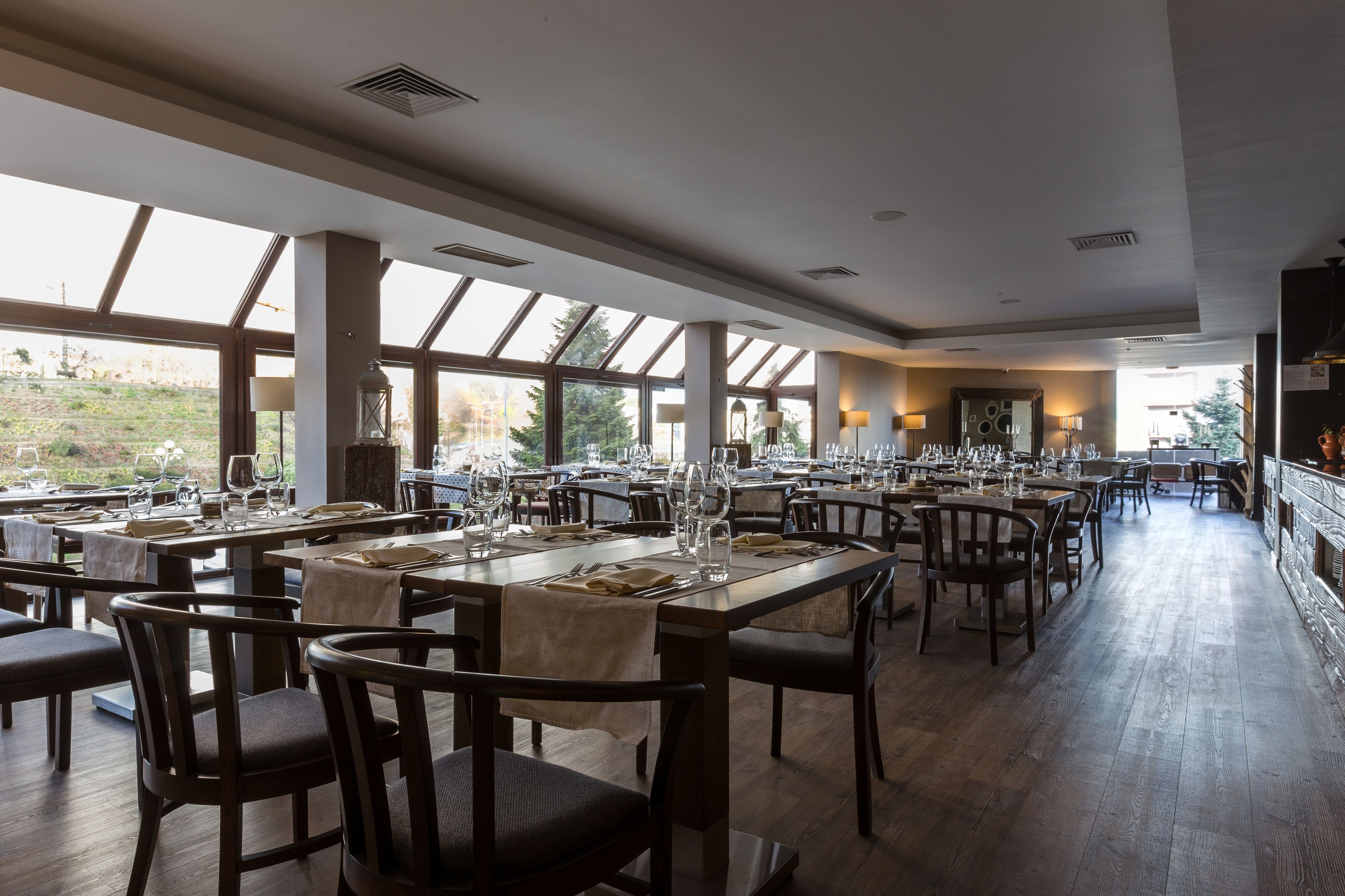 En rummelig restaurant med højt til loftet, store vinduer og trægulv. Bordene er dækket med hvide dugge og der er stole.