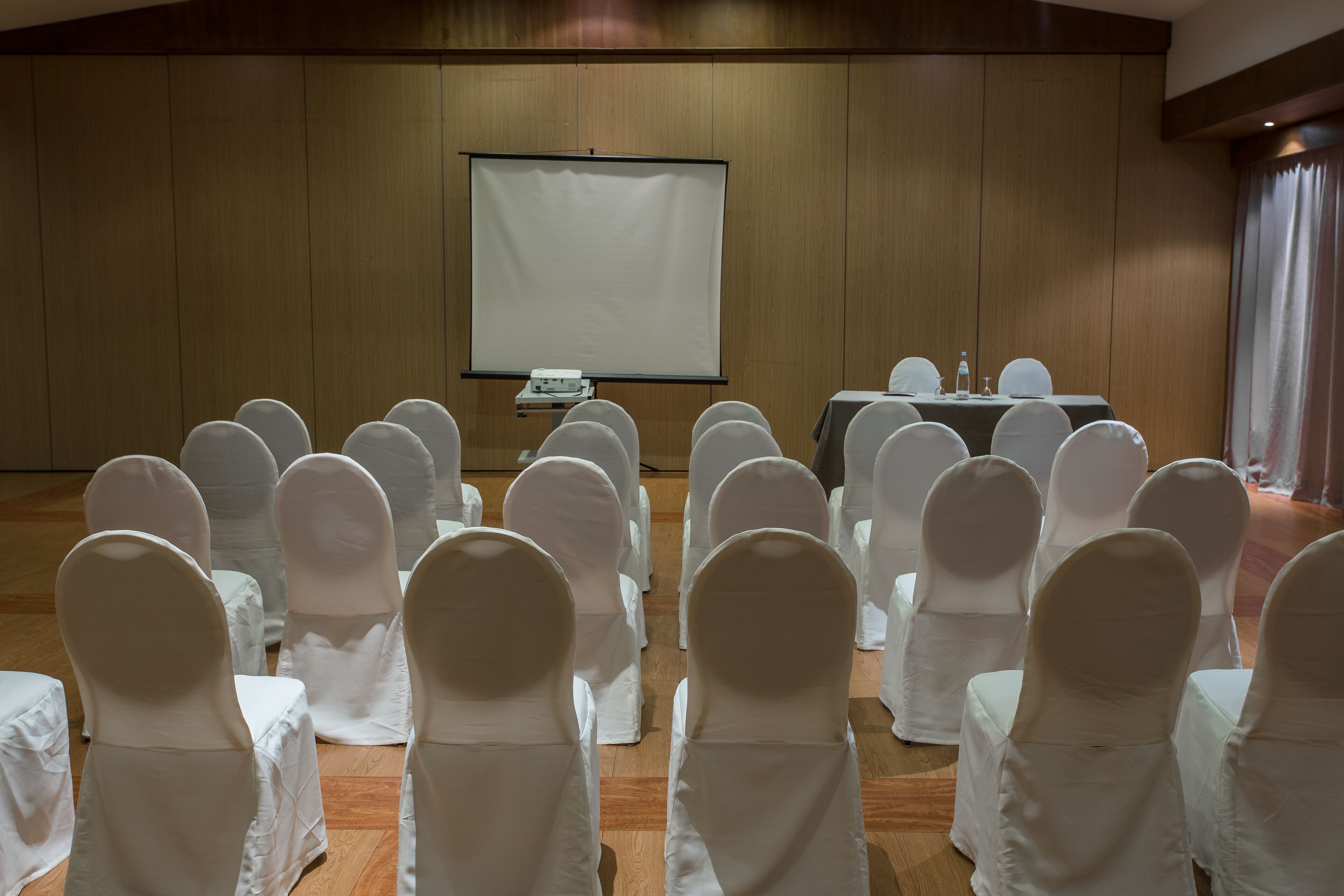 A conference room with rows of white-covered chairs facing a projection screen and a table with water bottles and glasses.