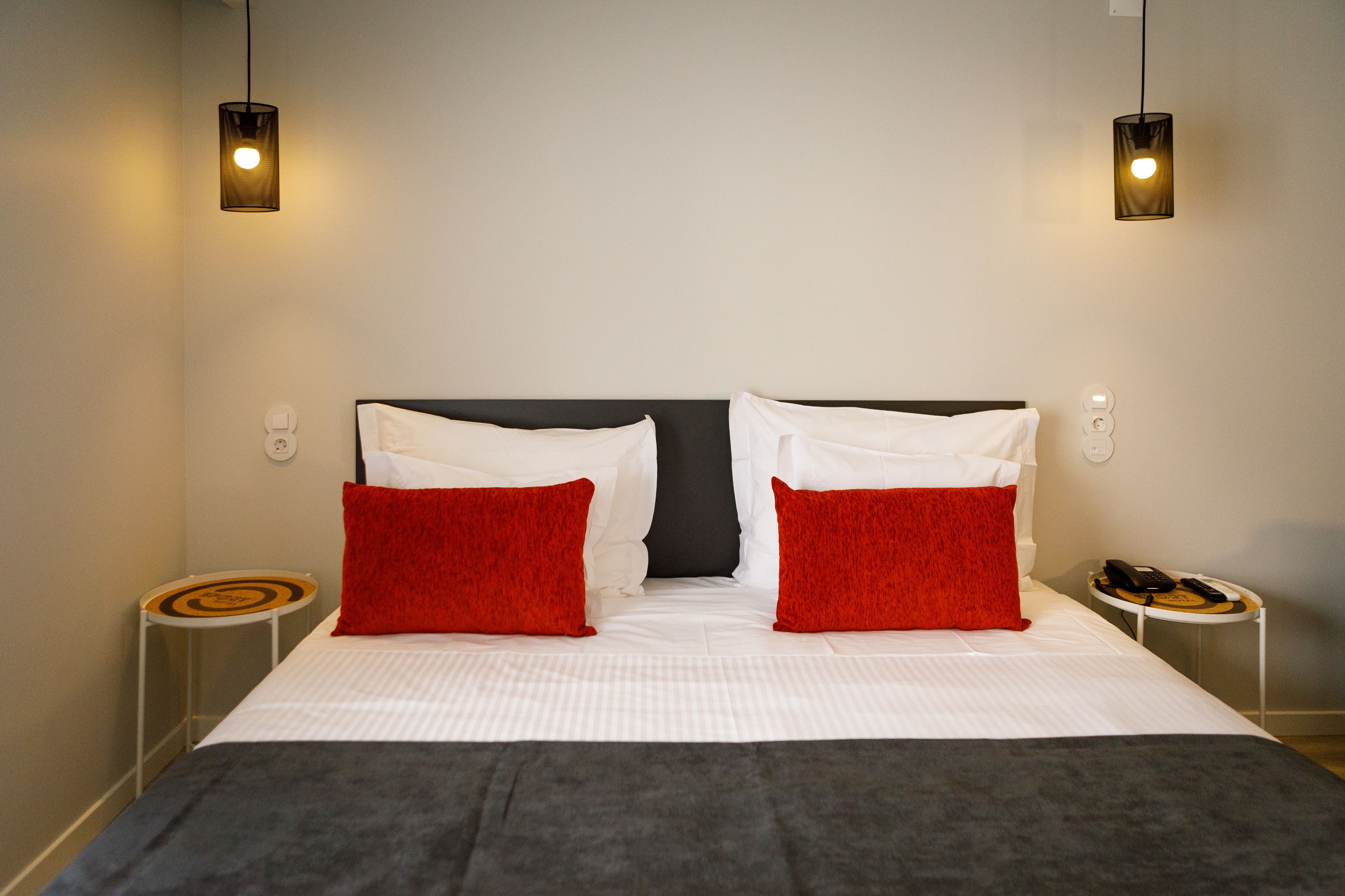 A neatly made bed with white and red pillows, two bedside tables with a phone and a book, and two wall-mounted lights.
