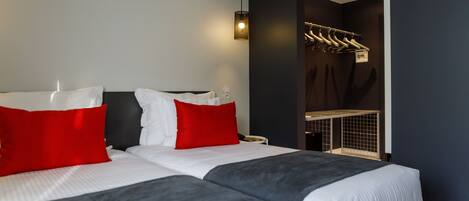 Una habitación de hotel moderna con dos camas, ropa de cama roja y blanca, un cabecero negro, un calentador de pared y un perchero.