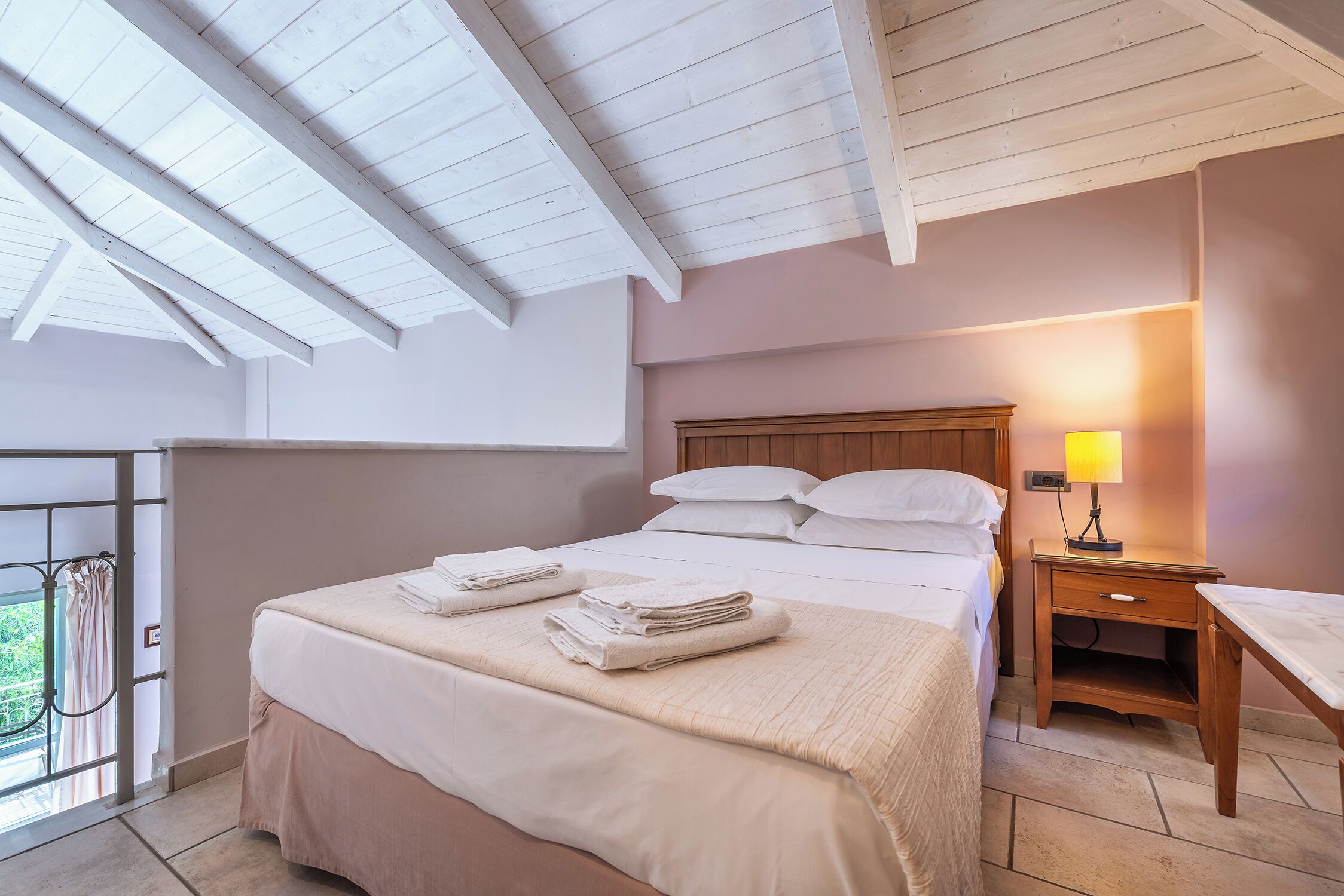 A bedroom with a bed, bedside table, and a window with a view of greenery.
