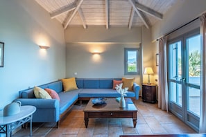 A living room with a blue sofa, a wooden coffee table, and a view through a large window. - Harmony Hotel Apartments (Aigialeia)