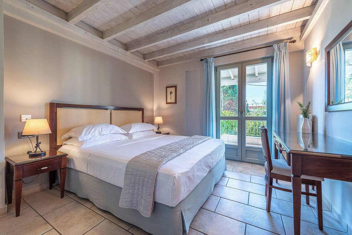 Quarto com uma cama grande, mesas de cabeceira com luminárias, uma escrivaninha e vista através de uma porta de vidro.