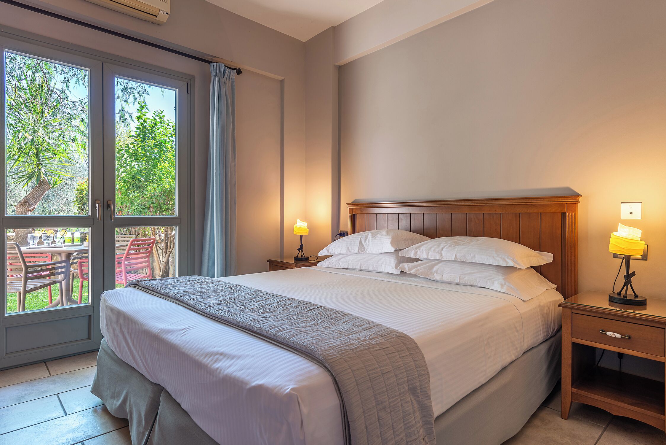 Quarto com uma cama grande, duas mesas de cabeceira com abajures, uma porta de vidro deslizante para um espaço de pátio e vista para o verde lá fora.