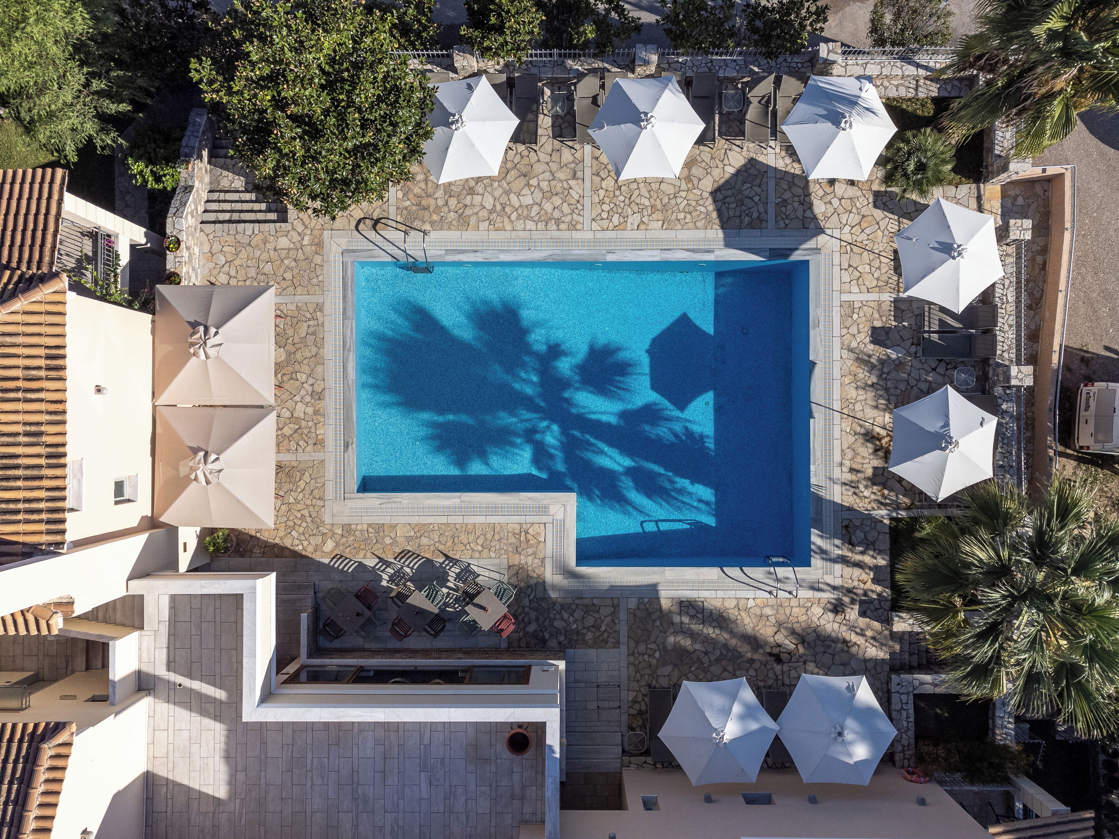 An aerial view of a swimming pool surrounded by lounge chairs and umbrellas, with stone walls and a tiled roof.