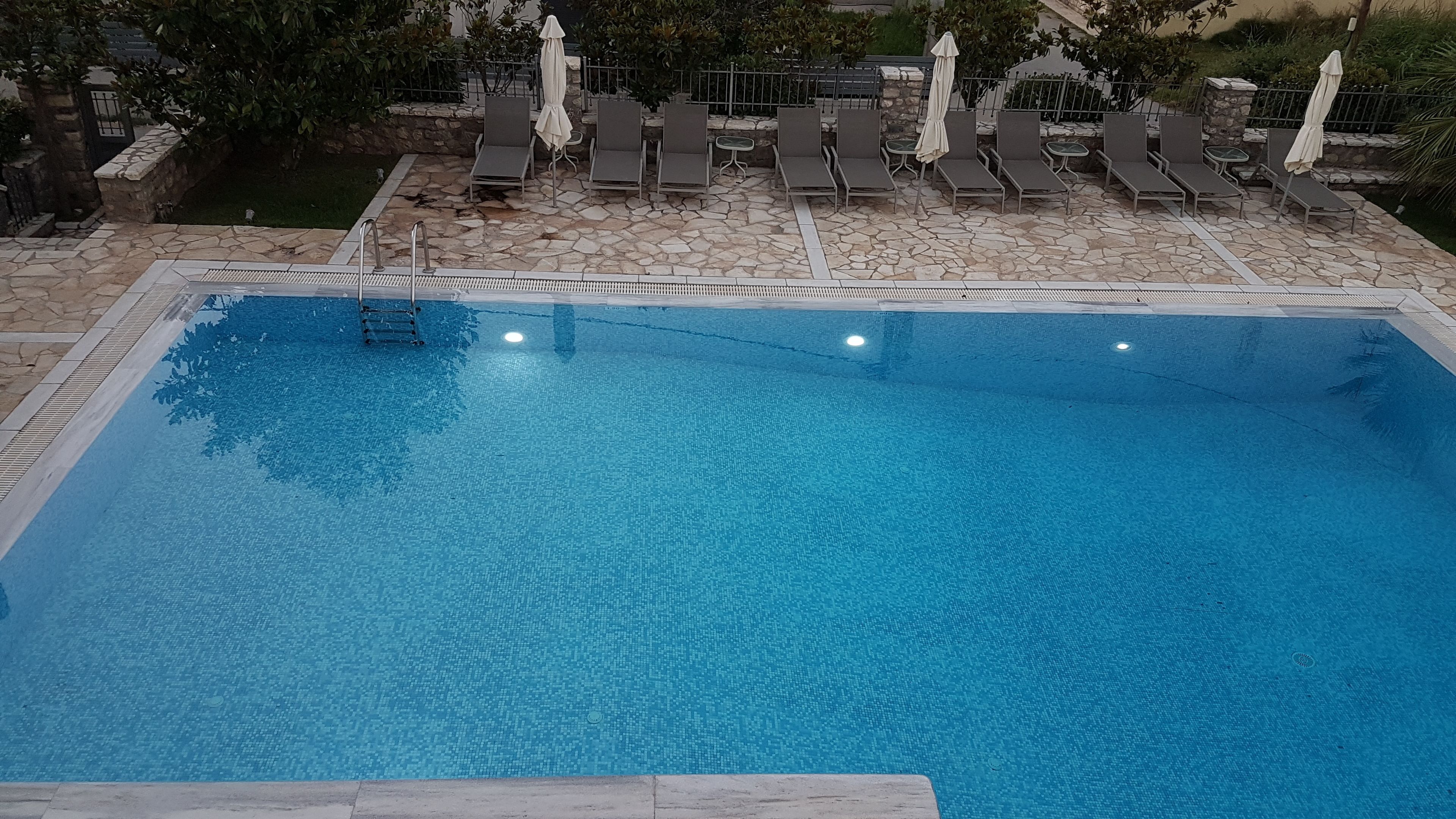 Una piscina con sillas de descanso y sombrillas, rodeada de baldosas de piedra y follaje.