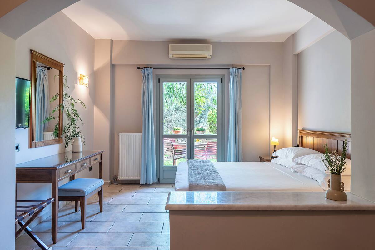 Quarto com cama, mesa de cabeceira e escrivaninha com cadeira. Há uma janela com cortinas, radiador e um vaso de planta.