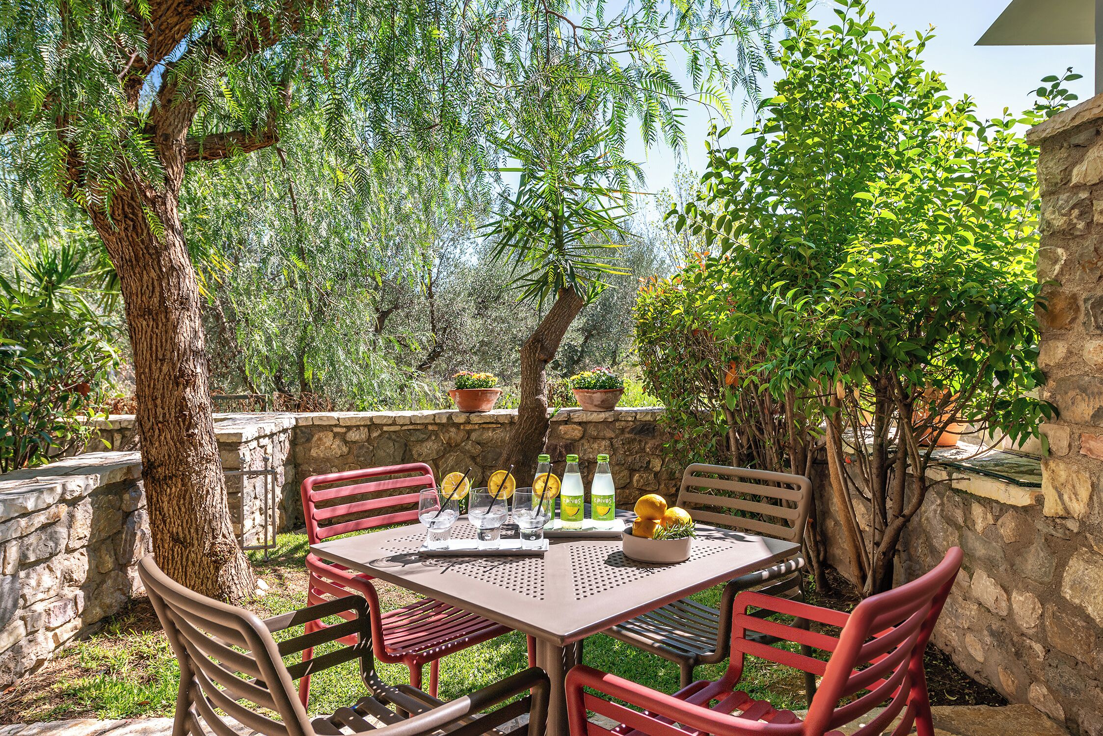 Un patio al aire libre con una mesa preparada para cuatro personas, rodeado de muros de piedra y abundante vegetación.