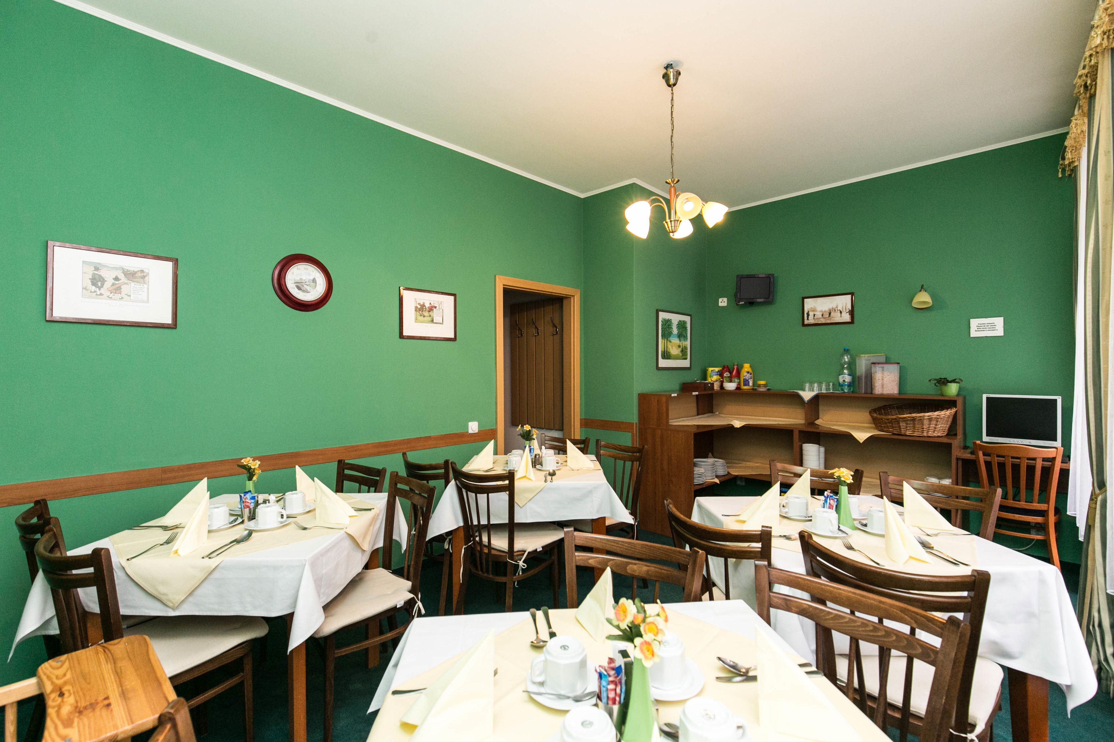 A dining area with green walls, wooden tables, and chairs. There are framed pictures on the walls, a clock, and a television on a shelf.