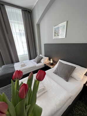 A hotel room with two beds, a nightstand, a bedside lamp, a framed picture on the wall, and a window with curtains. - Hotel Boston (Karlovy Vary)