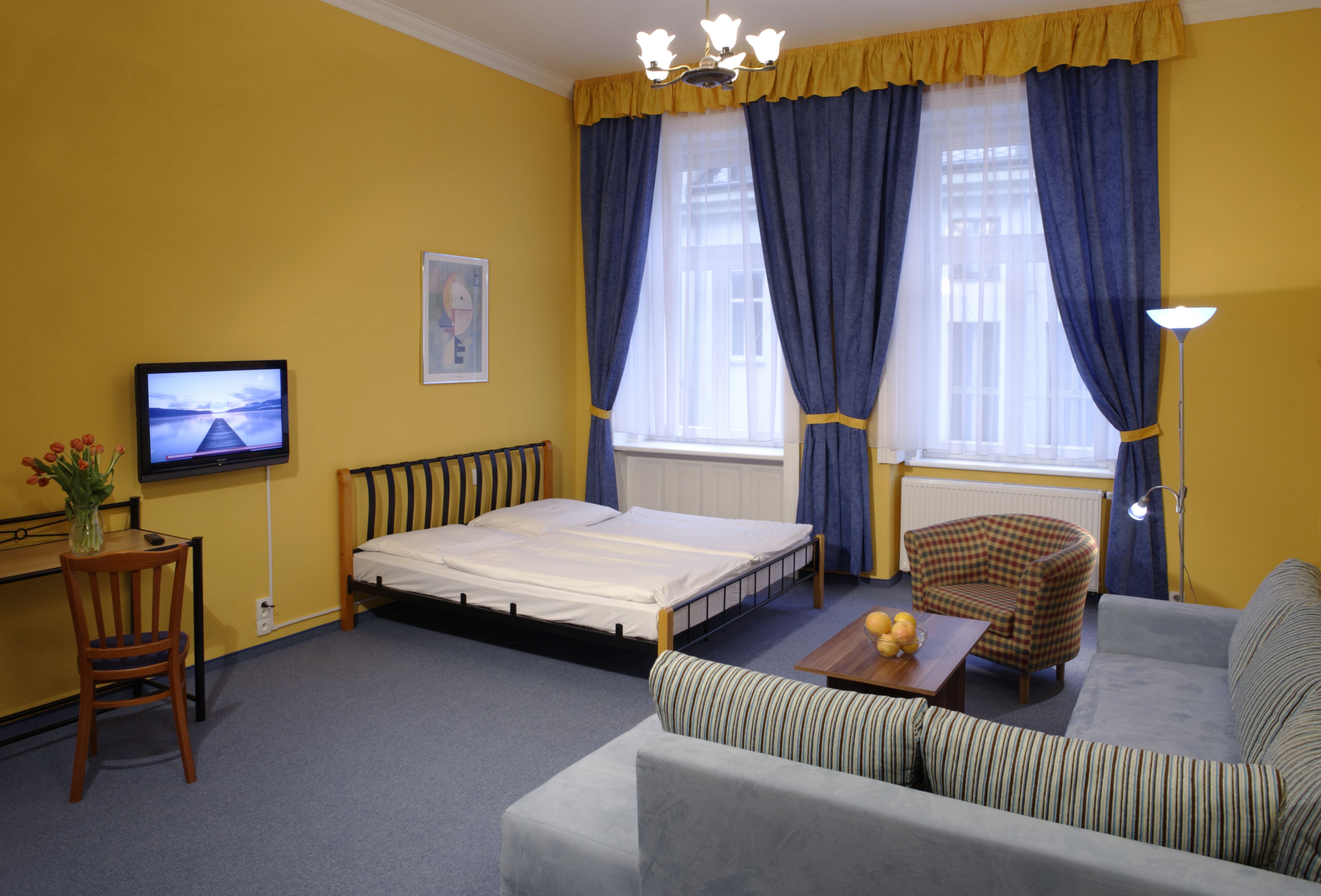 A hotel room with a bed, a sofa, a chair, a desk with a computer, and a TV on the wall.