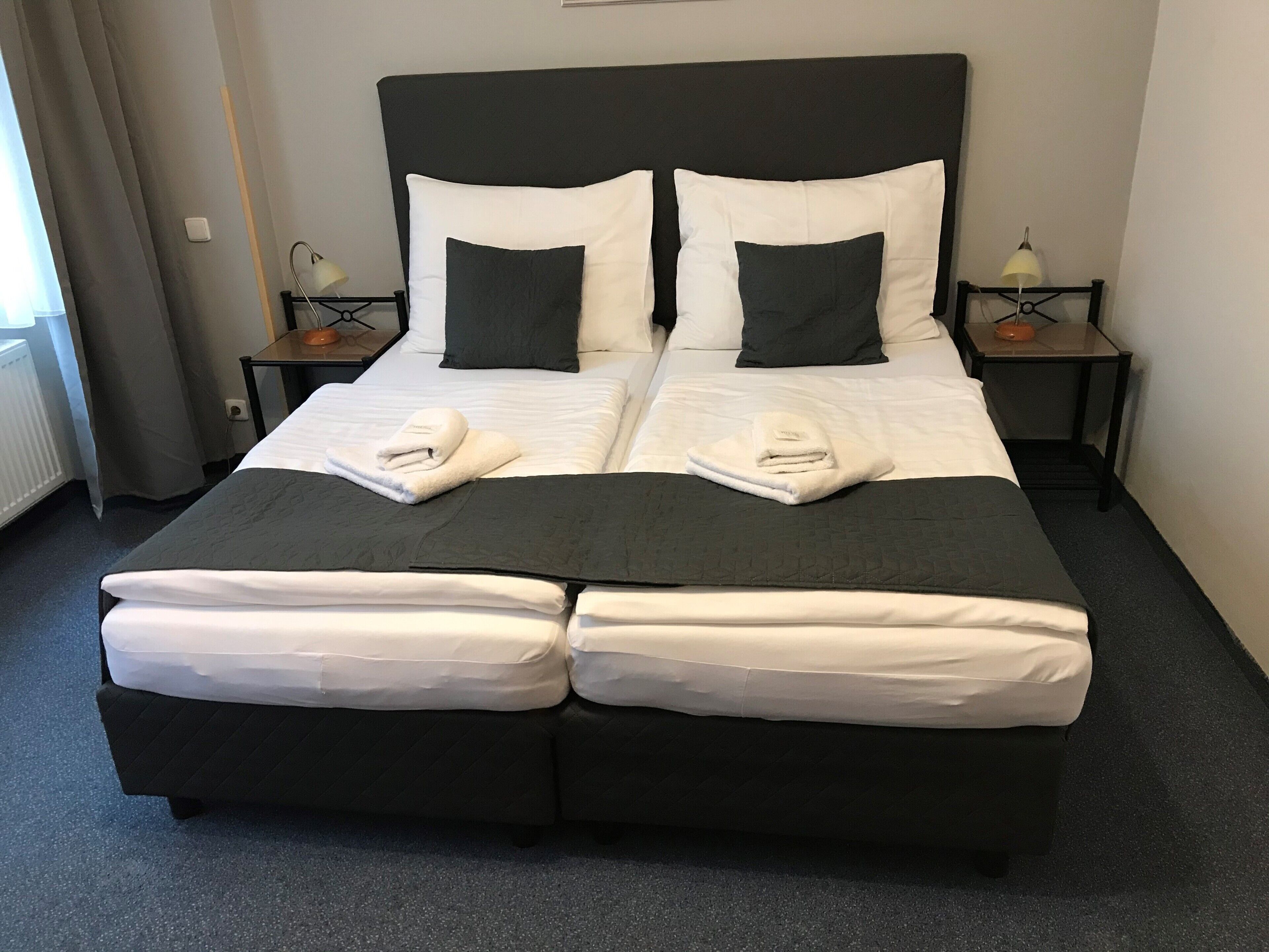 Cama de casal com duas almofadas, cabeceira preta e edredom branco. Há duas mesas de cabeceira com luminárias e uma janela com cortinas.