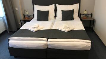 Cama de casal com duas almofadas, cabeceira preta e edredom branco. Há duas mesas de cabeceira com luminárias e uma janela com cortinas.